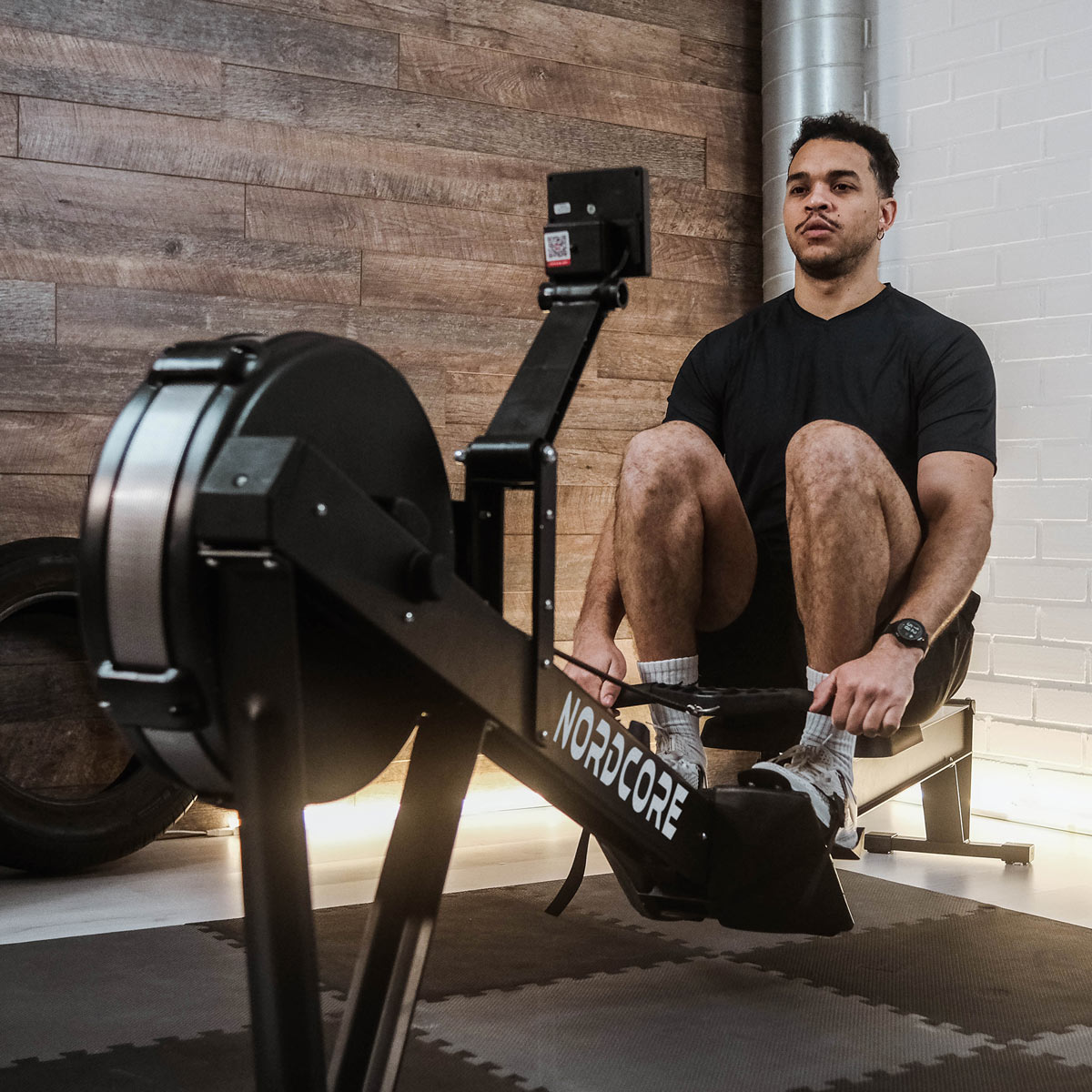 Ein Mann in schwarzem T-Shirt und Shorts sitzt auf einem schwarzen Rudergerät und stellt die Fußschlaufen in einem Fitnessstudio ein.