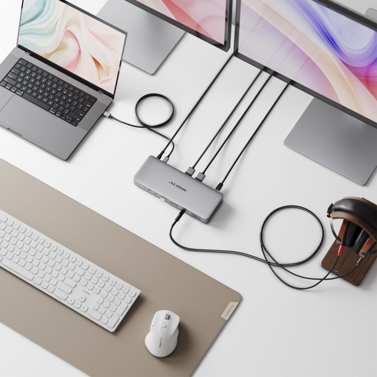 Ein grauer Laptop, eine graue Dockingstation, zwei Monitore, eine weiße Tastatur und Maus auf einem weißen Schreibtisch mit beiger Matte.