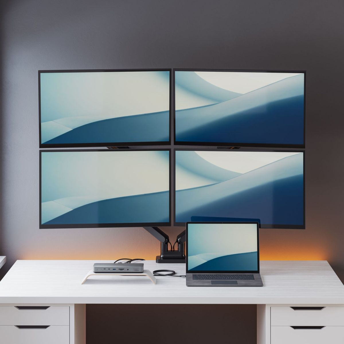 Vier Monitore über einem Schreibtisch mit Laptop und Dockingstation.