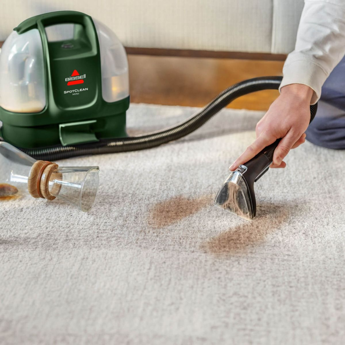 Eine Person benutzt einen grünen Bissell SpotClean Mini Teppichreiniger, um eine braune Verschüttung von einem hellgrauen Teppich zu entfernen.