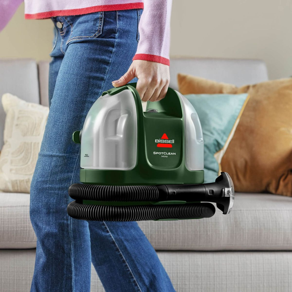 Eine Person in blauen Jeans und einem pinkfarbenen Oberteil hält einen grünen und durchsichtigen Bissell SpotClean Mini tragbaren Teppichreiniger.