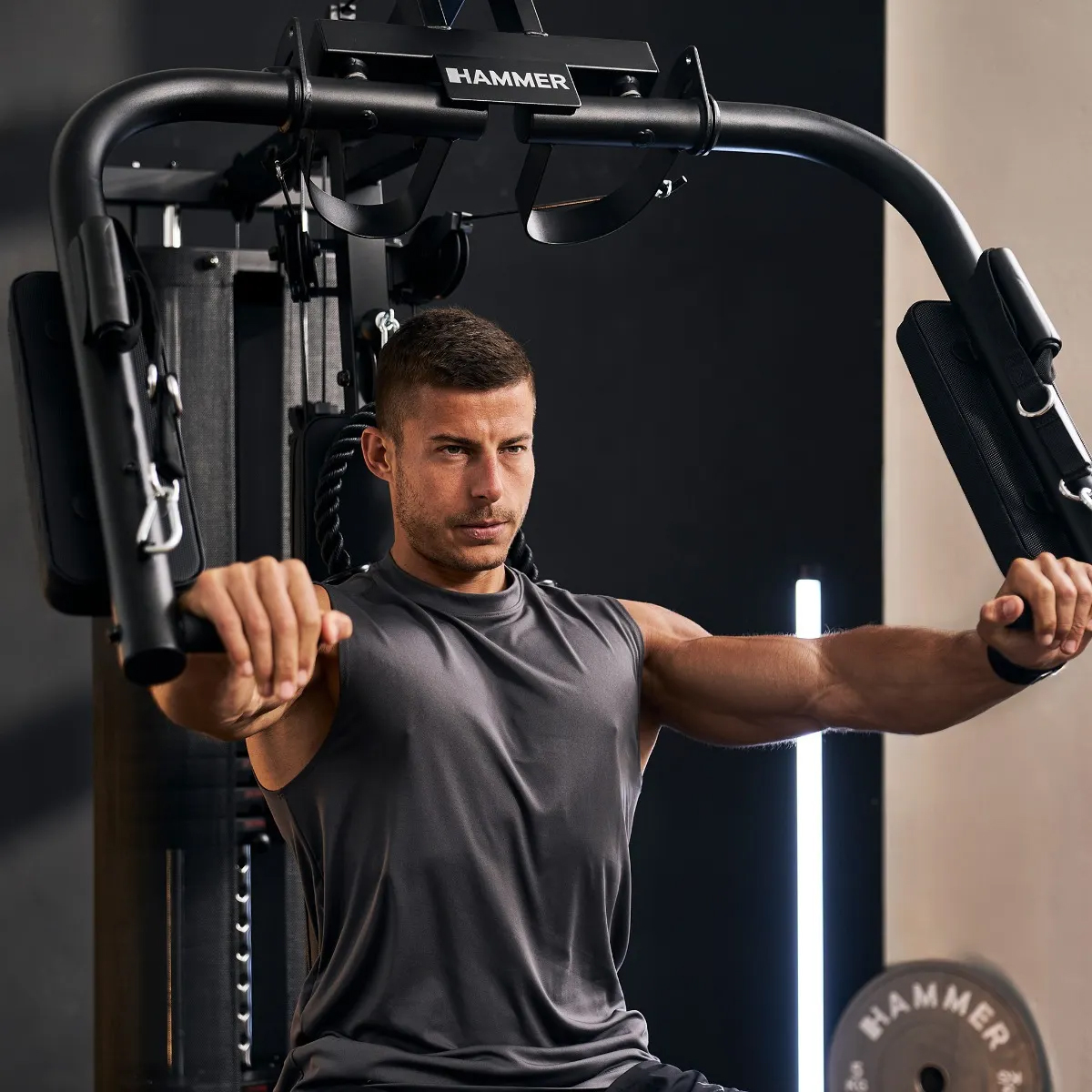 Ein Mann in einem grauen Tanktop trainiert konzentriert an einer schwarzen Hammer-Brustpresse in einem Fitnessstudio.