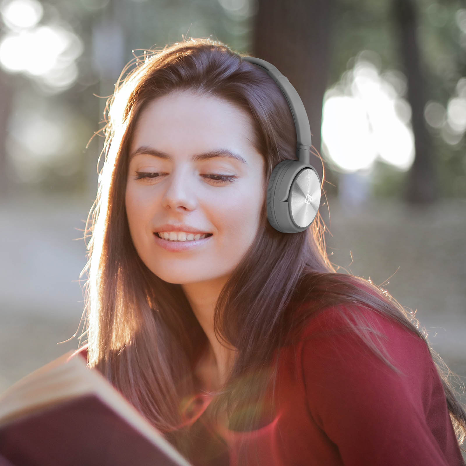 Woman with headphones reads a book outdoors in the sunlight.