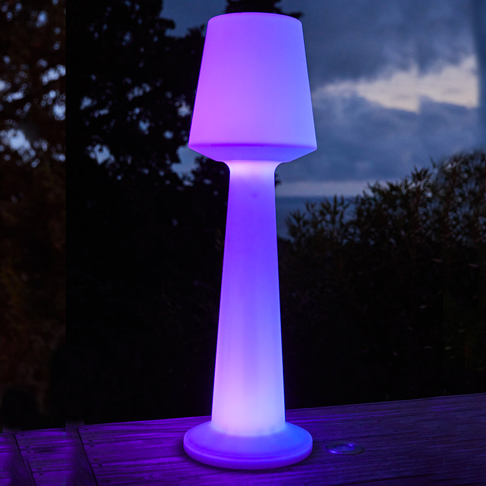 Eine beleuchtete Stehlampe in Lila und Blau erhellt einen Holzboden im Freien bei Dämmerung.