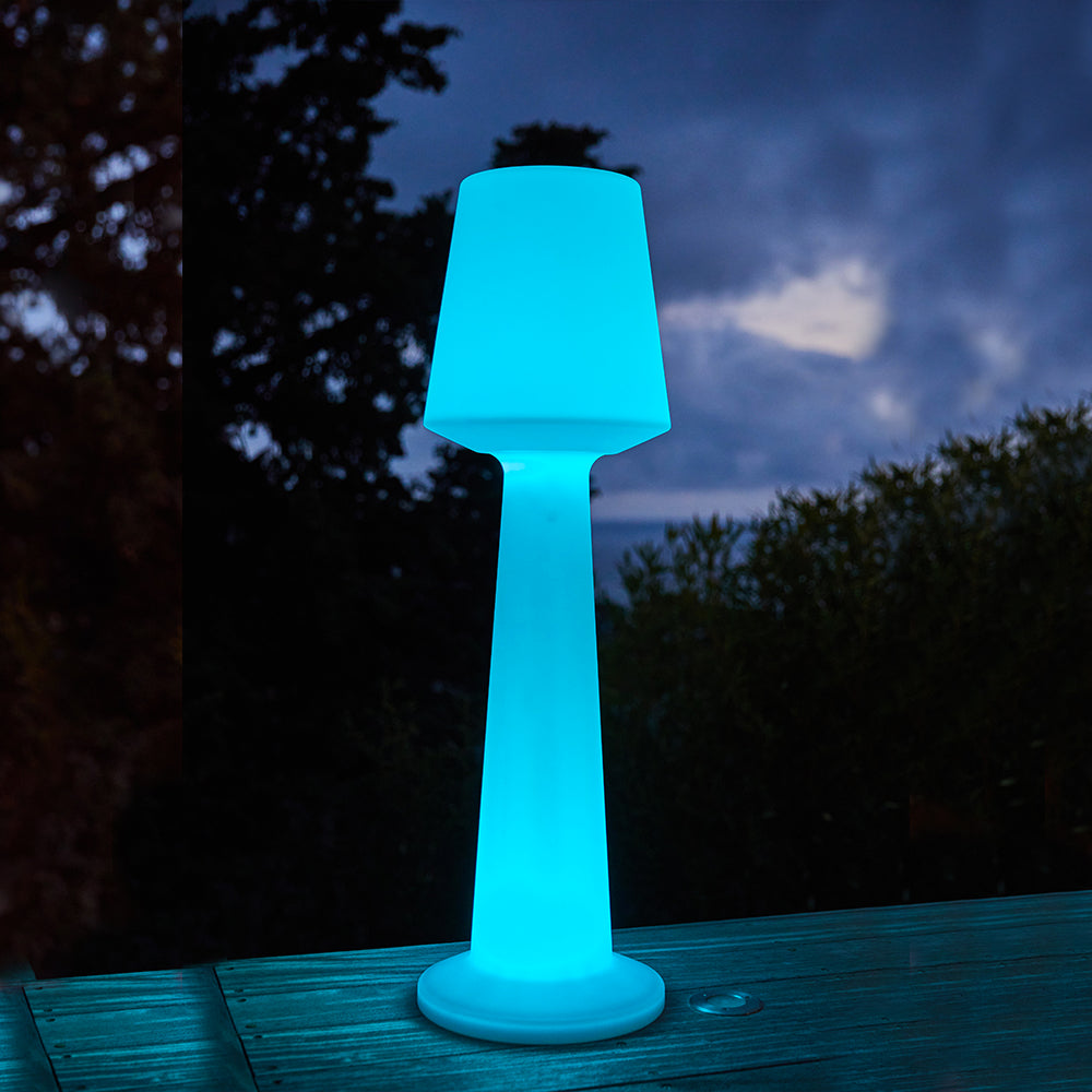 Eine leuchtende blaue Stehlampe steht auf einem Holzboden im Freien bei Dämmerung.