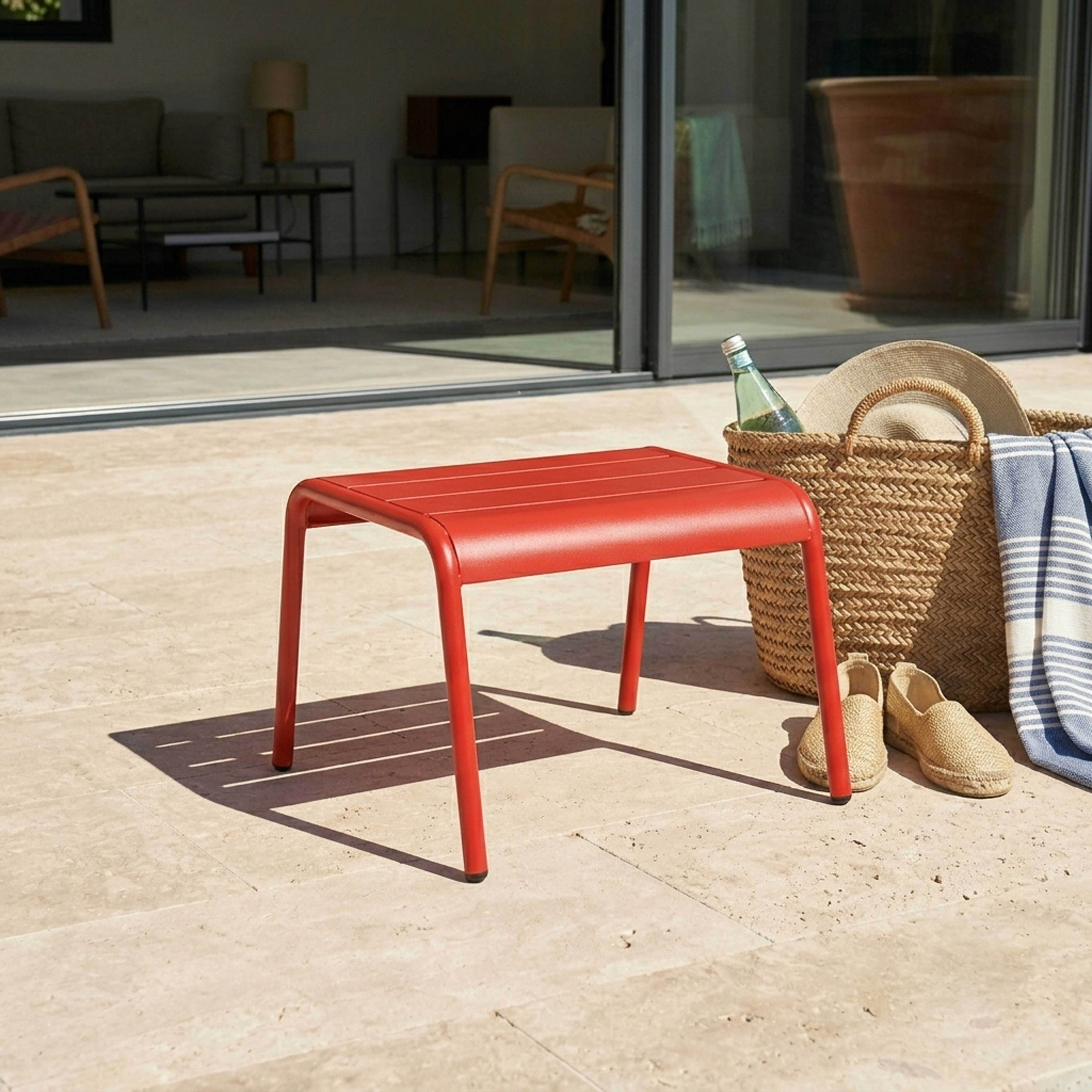 Ein roter Hocker steht auf einer beigefarbenen Terrasse neben einem Korbflechtkorb.