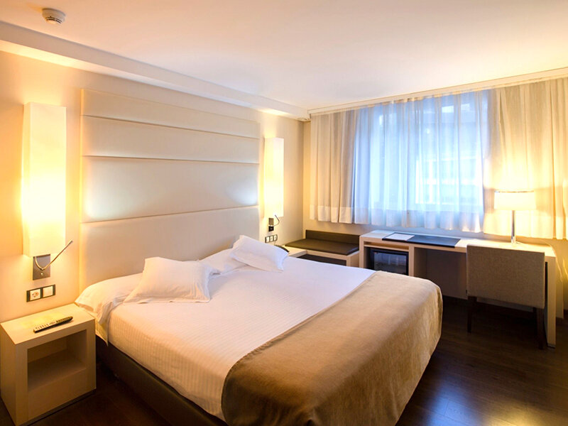 Una habitación de hotel con cama grande, mesita de noche, escritorio, silla y ventana con cortinas.