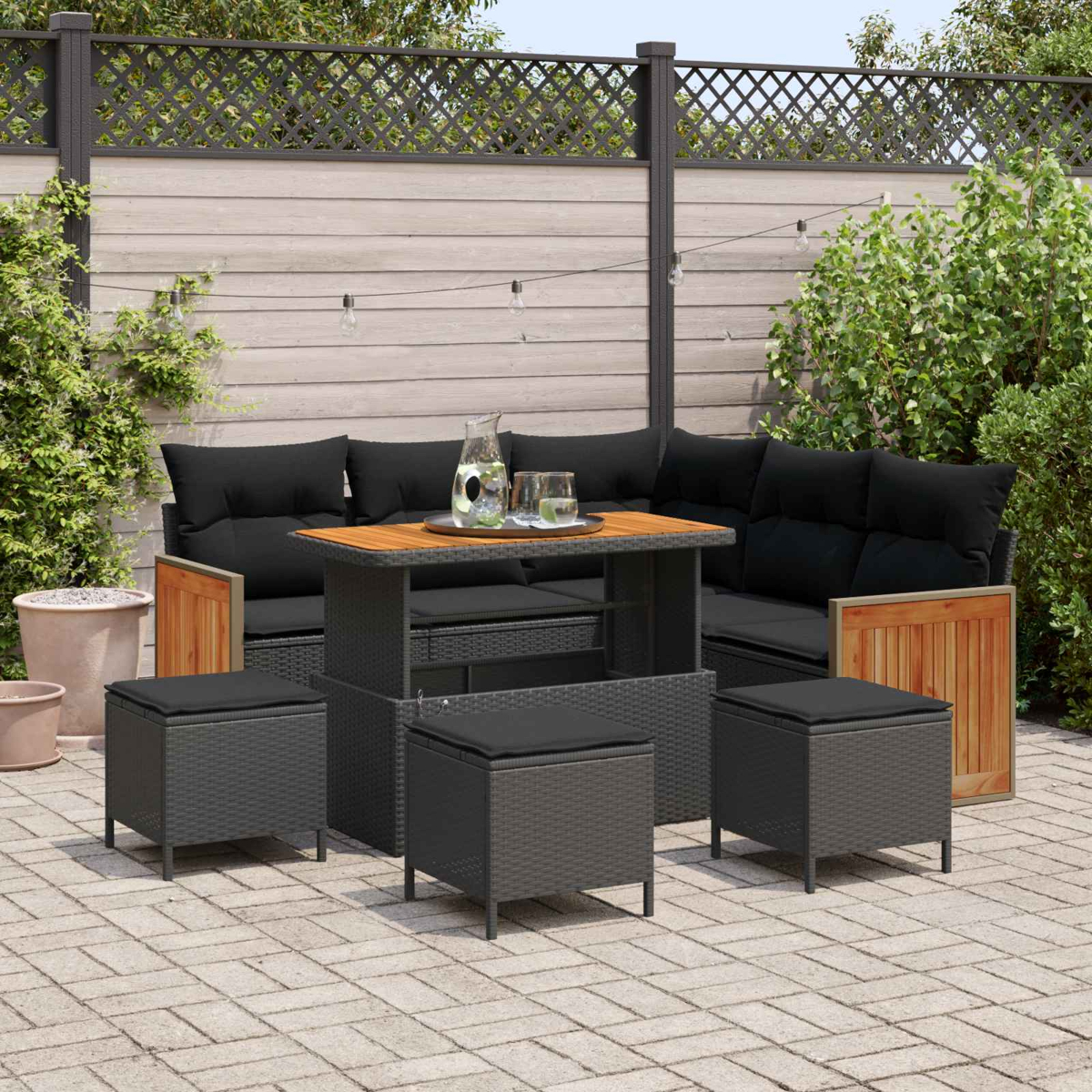 Gartenlounge mit schwarzem Ecksofa, Tisch und Hockern auf einer gepflasterten Terrasse.