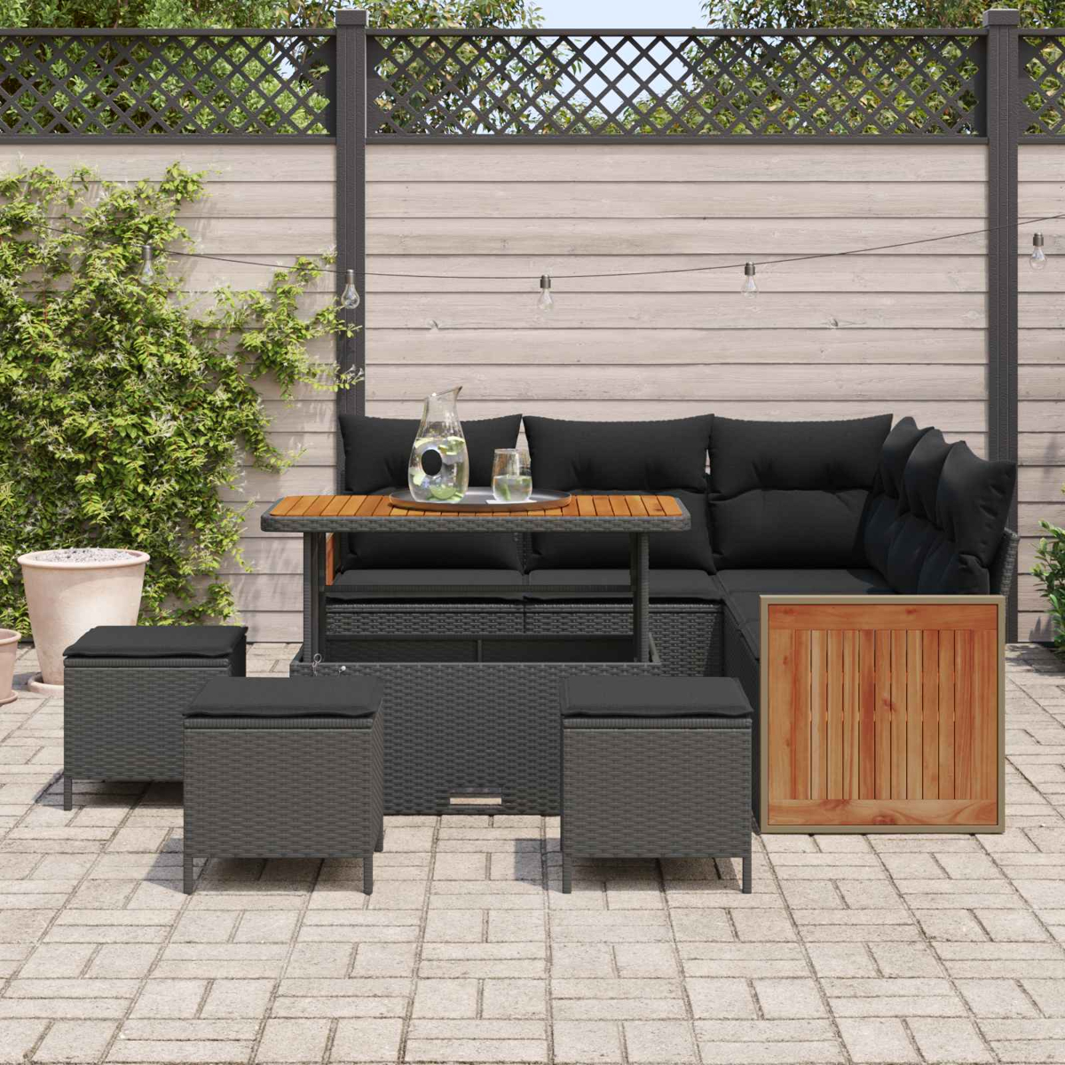 Ein dunkles Rattan-Loungeset mit schwarzen Kissen und einem Holztisch auf einer Kopfsteinpflasterterrasse.