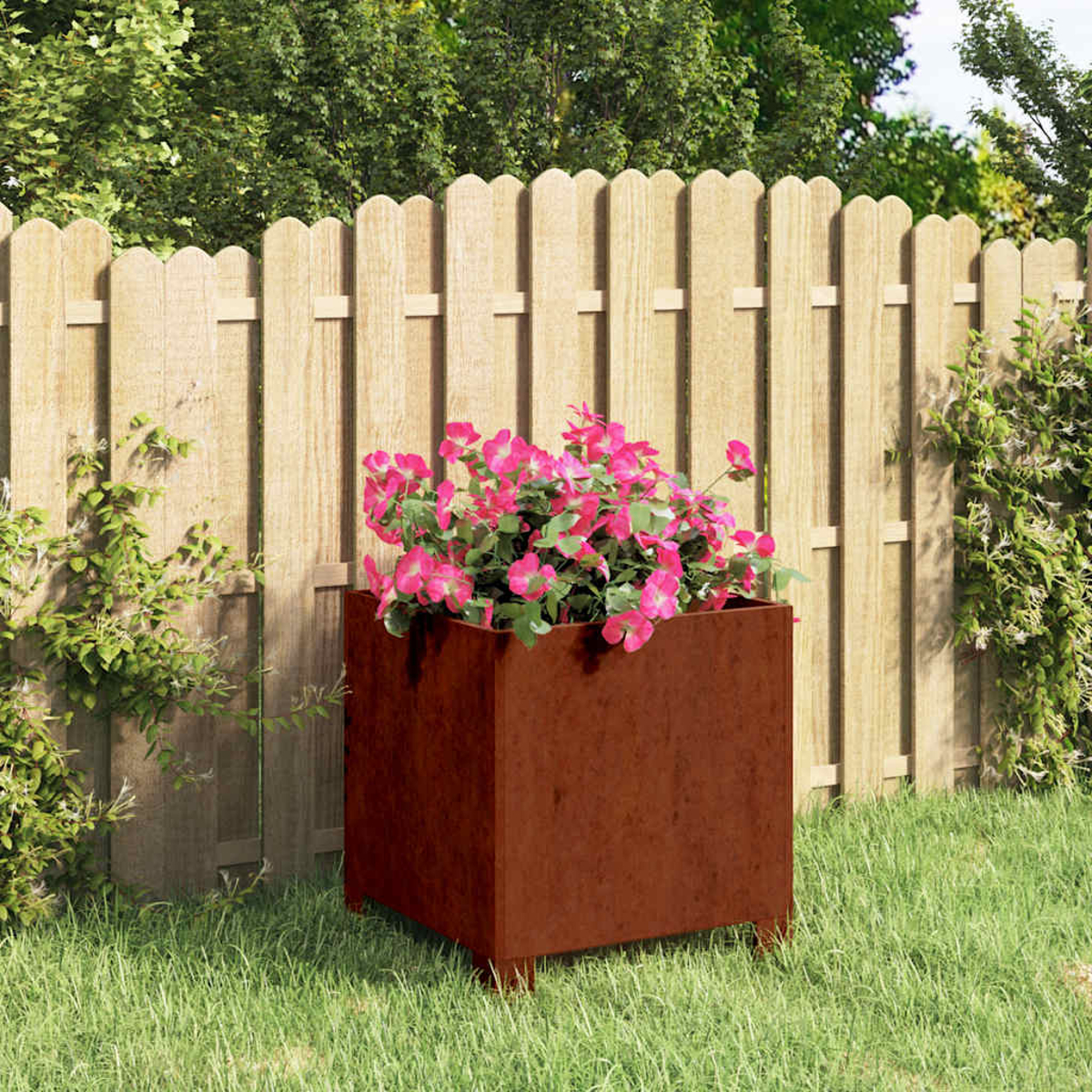 Ein rostbrauner Blumenkübel mit rosa Blumen steht im Gras vor einem Holzzaun.