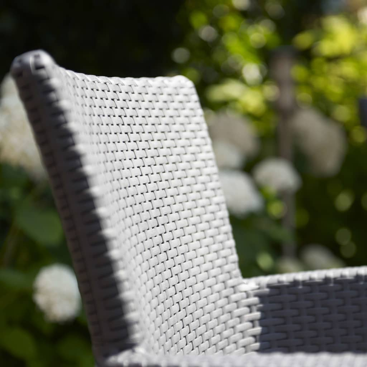 Una sedia in rattan grigio in un giardino con fiori bianchi sfocati.