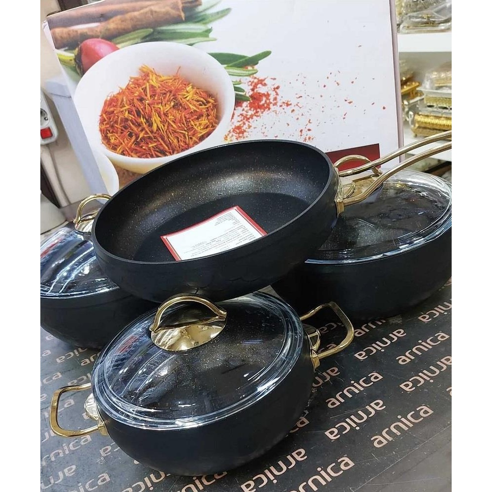Altın kulplu ve cam kapaklı siyah tencere seti, üzerinde 'arnica' baskısı olan masa örtüsünde duruyor.