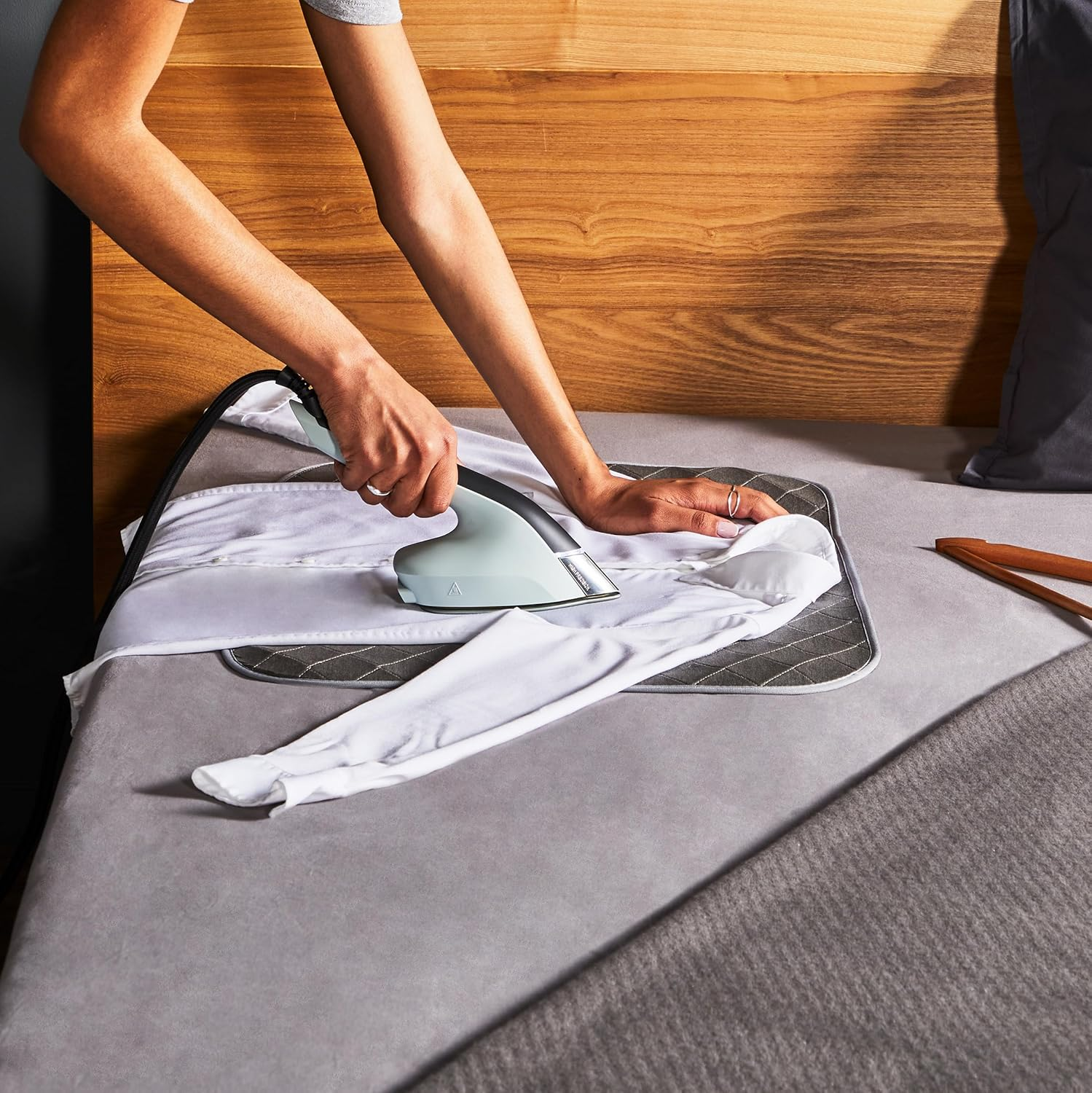 Person bügelt ein weißes Hemd auf einem Bügelbrett auf einem Bett.