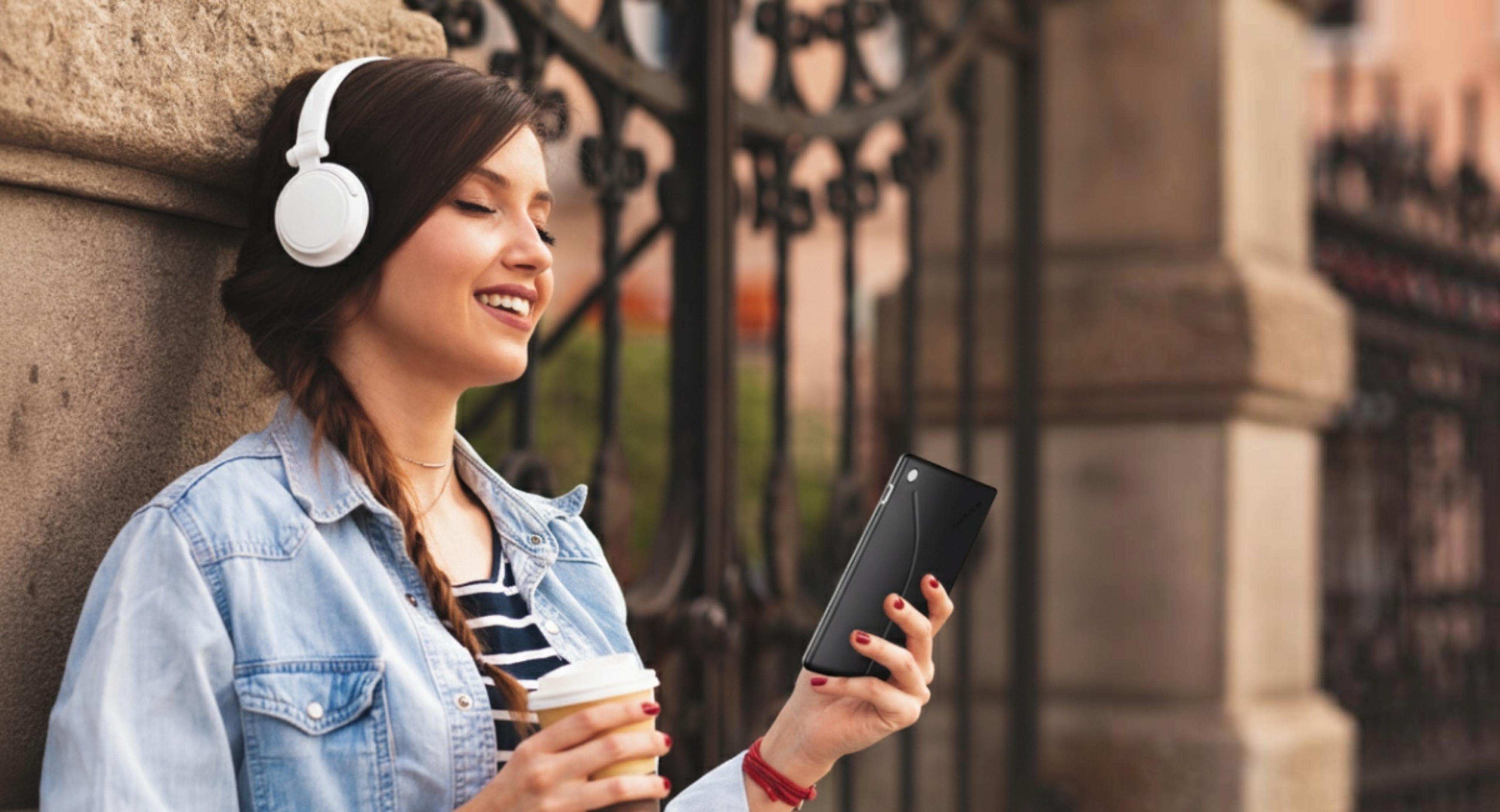 Una donna con cuffie e tazza di caffè, tiene in mano uno smartphone.
