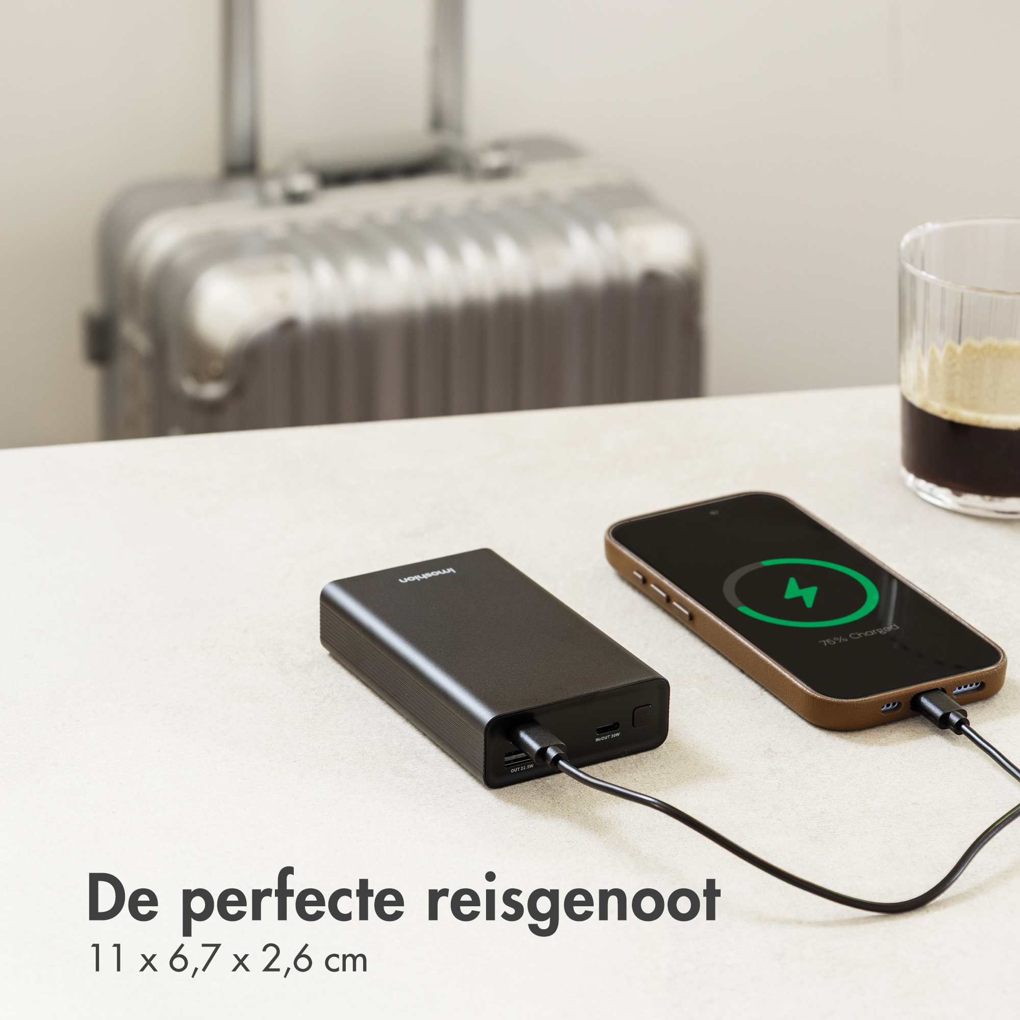 Power bank nera carica uno smartphone. Valigia e bicchiere di caffè sullo sfondo.