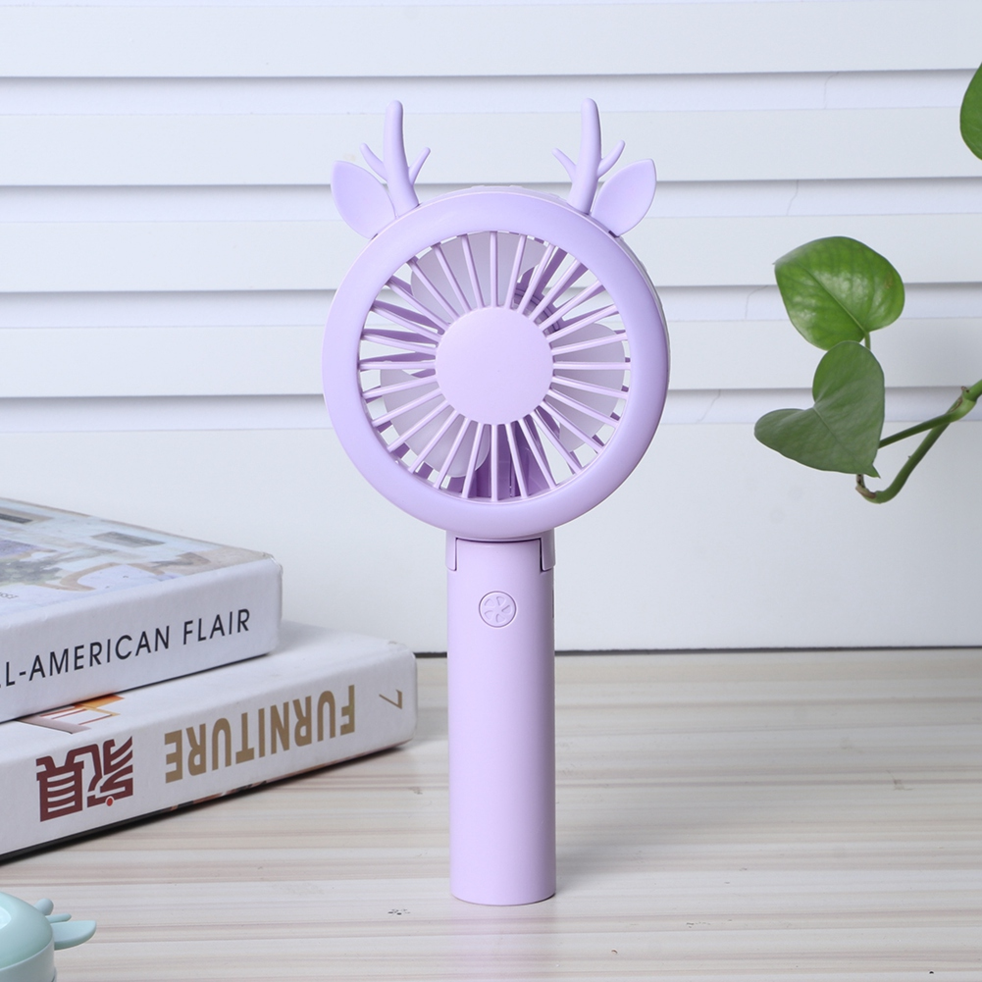 Ein lila tragbarer Ventilator in Hirschform mit Flügeln steht neben Büchern auf einem Tisch.