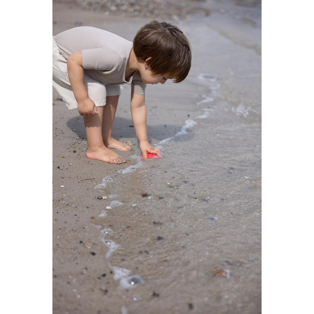 Ein Kind in beiger Kleidung spielt mit einem roten Spielzeug an einem Sandstrand mit sanften Wellen.