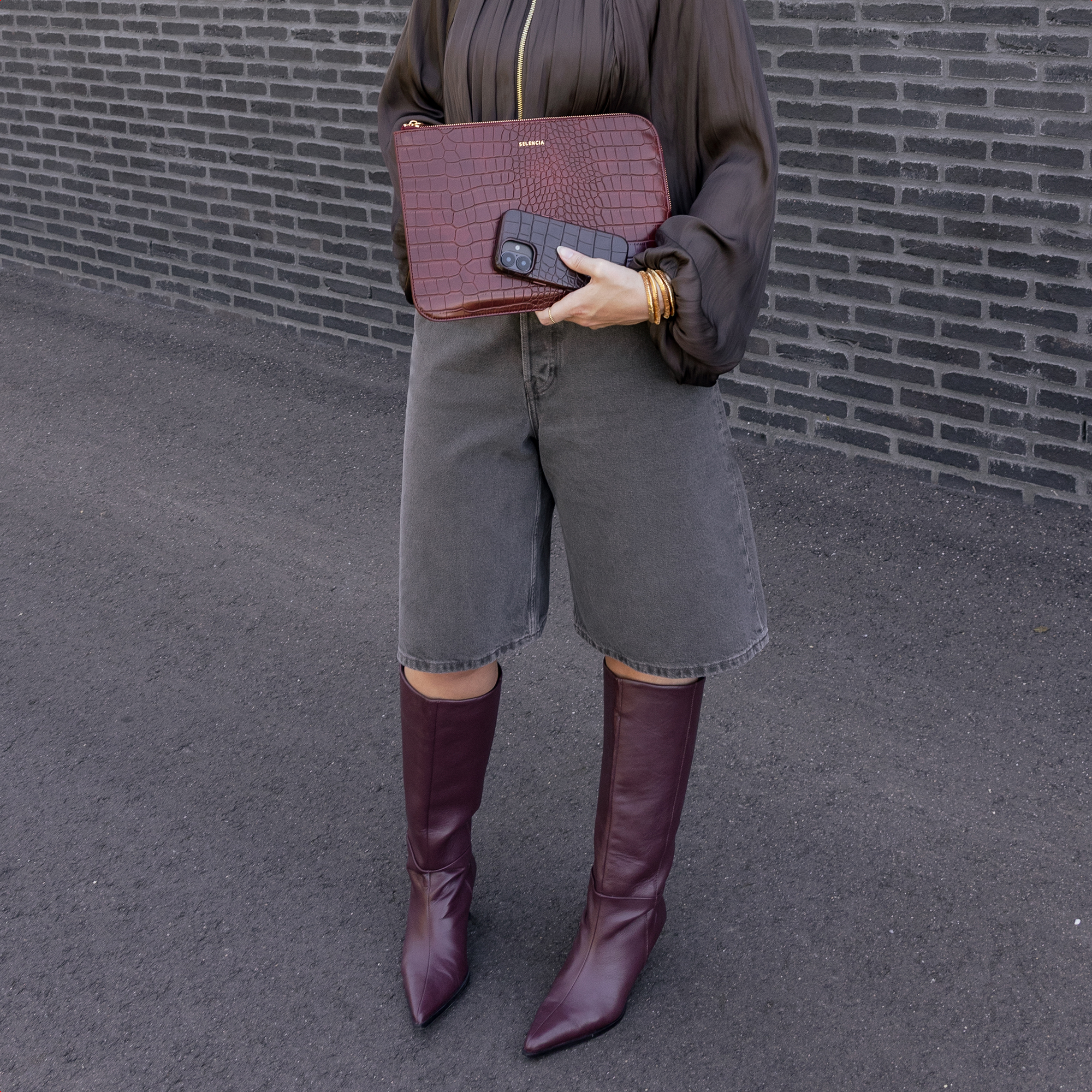 Frau mit dunkelroter Kroko-Tasche, Handy und Stiefeln vor Ziegelwand.