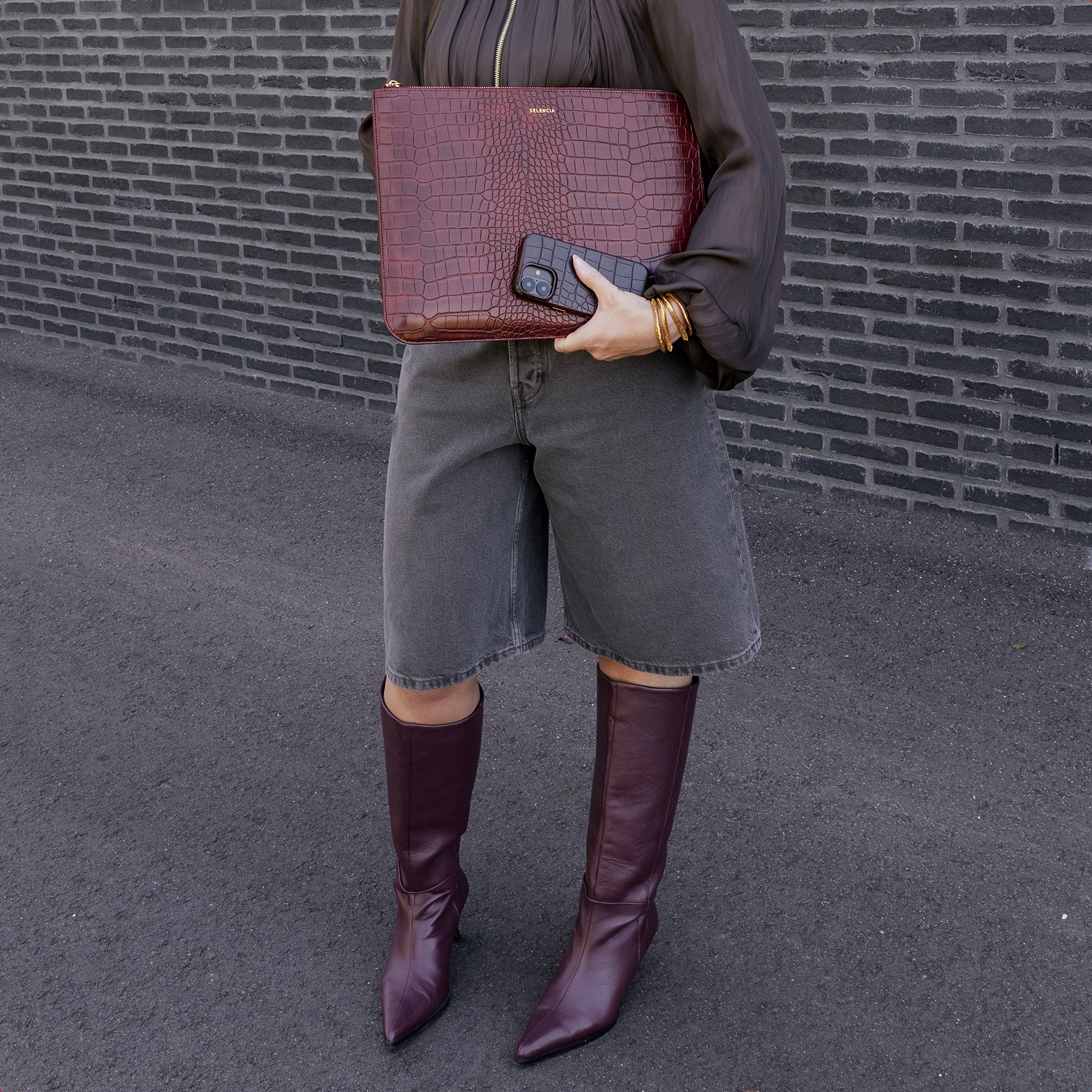 Eine Person hält eine burgunderrote Clutch mit Alligatorprägung und ein Handy. Sie trägt Jeansshorts und burgunderrote Stiefel.