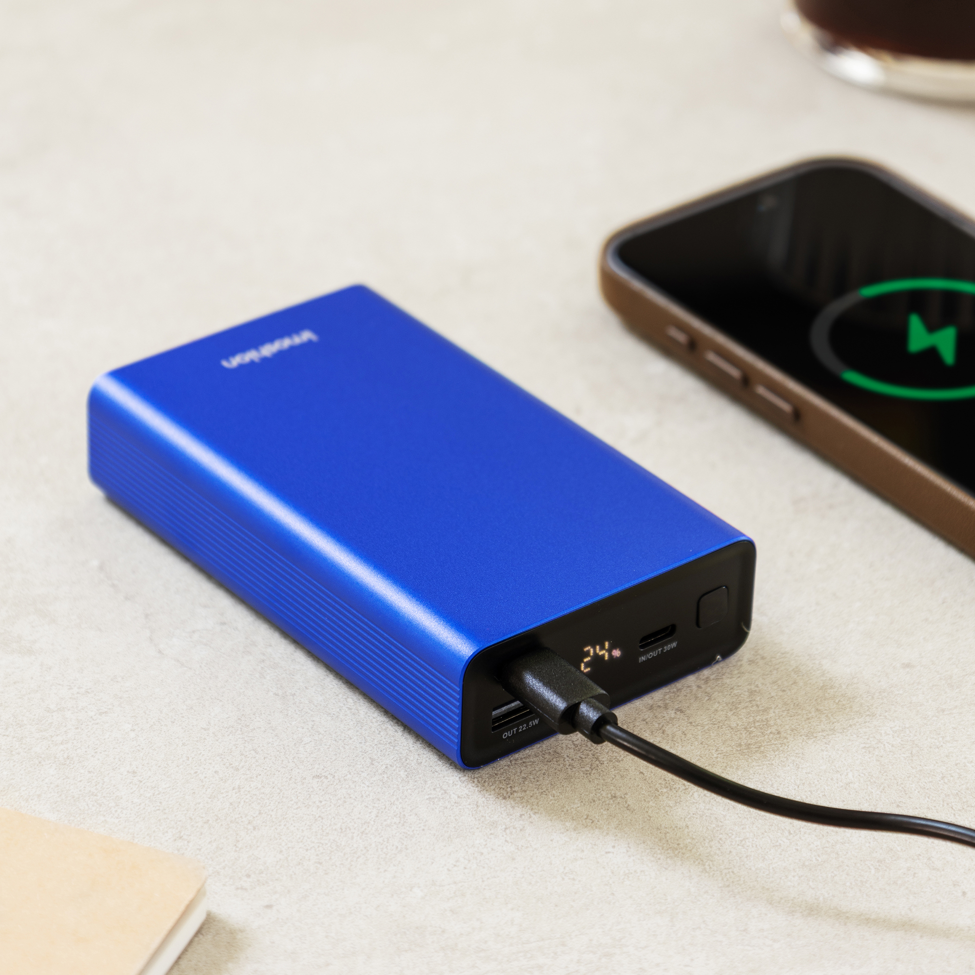A blue power bank is charging a phone, with a coffee cup in the background.