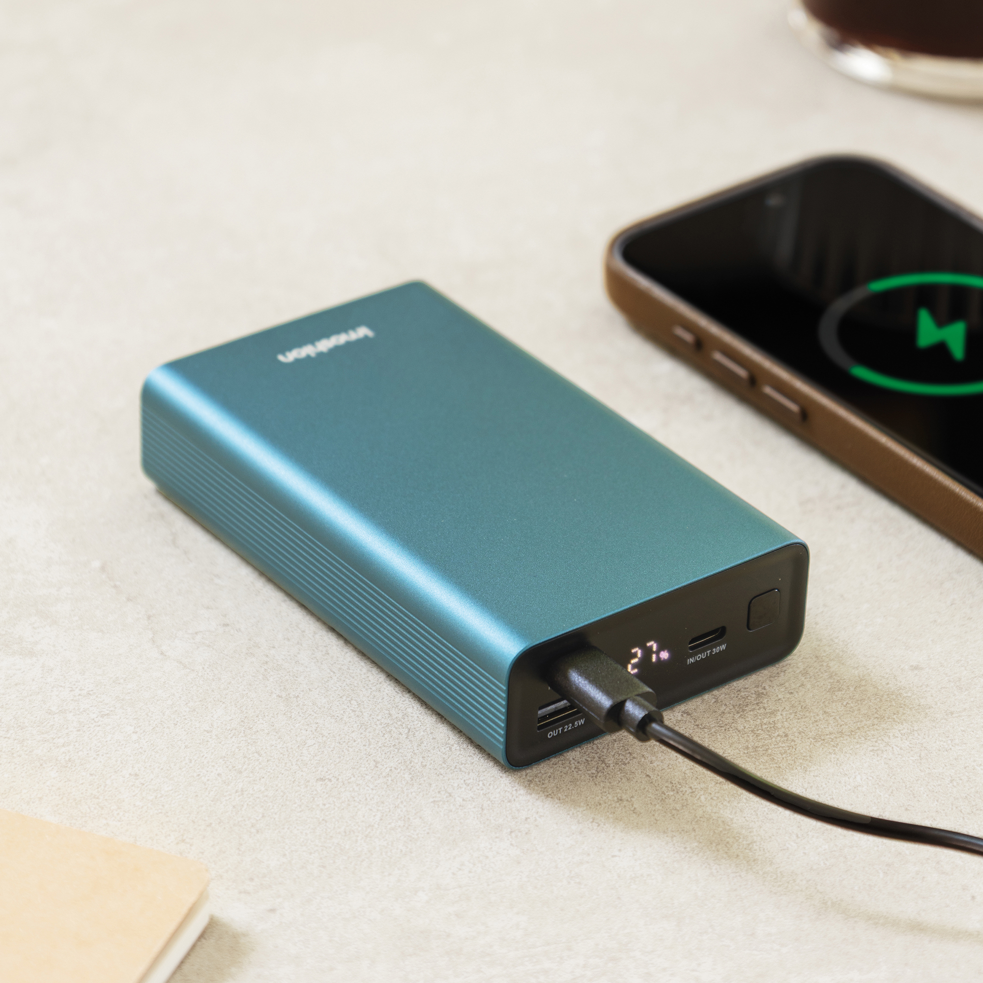 Un power bank color verde acqua con display digitale che mostra il 27% è collegato a uno smartphone.