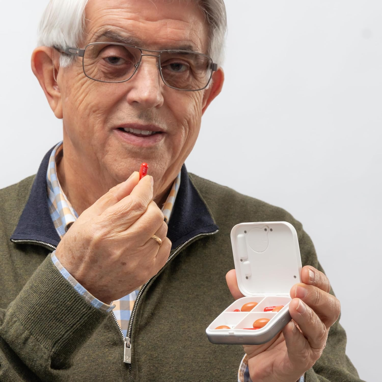 Ein älterer Mann mit Brille hält eine rote Pille und eine Pillendose.
