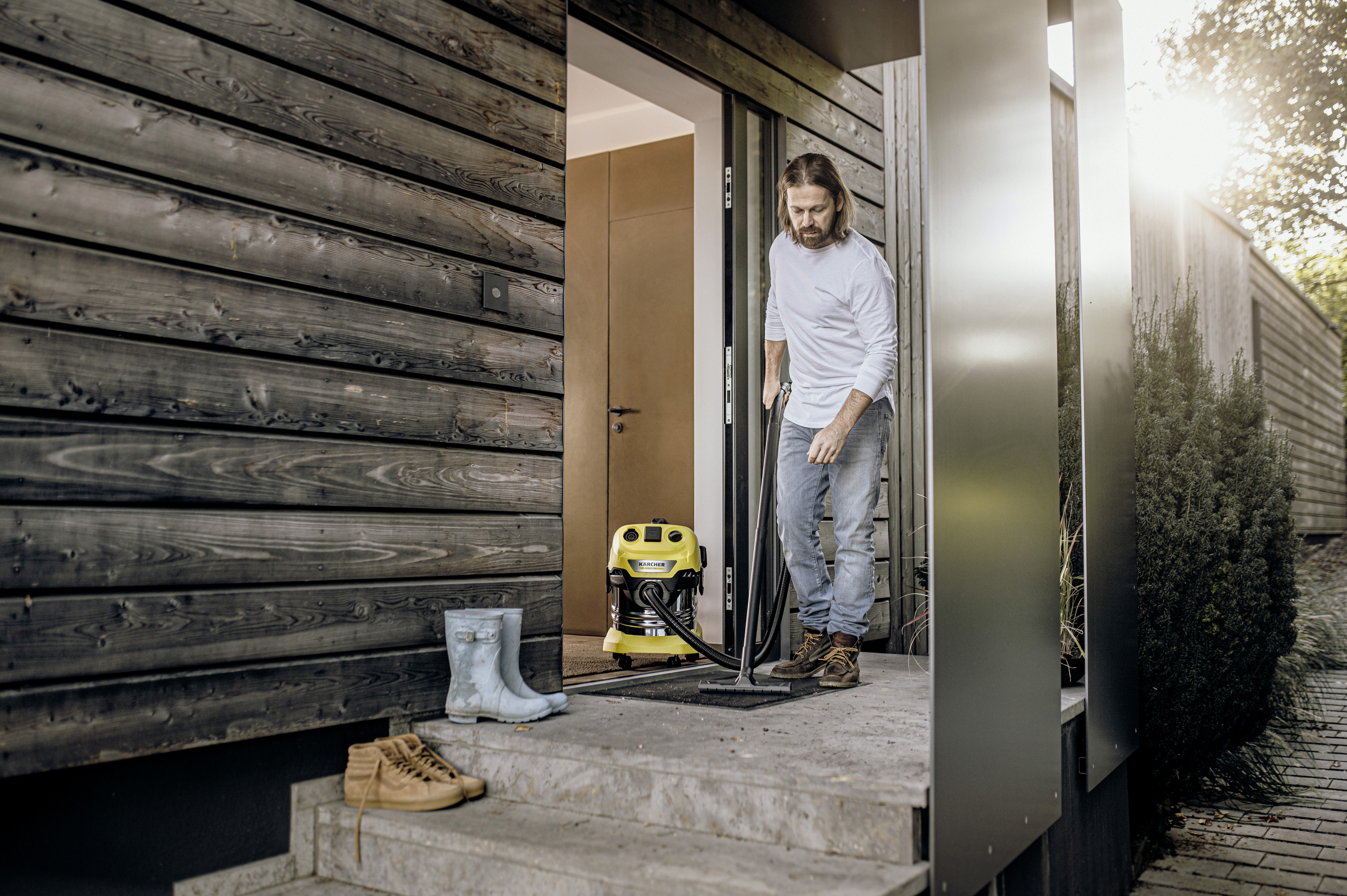 Uomo aspira con aspirapolvere Karcher giallo davanti a case di legno. Stivali e scarpe vicini.