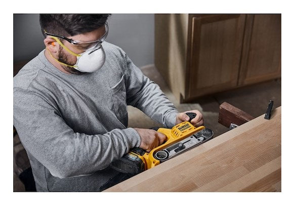 Uomo con occhiali e maschera leviga legno con levigatrice a nastro gialla.