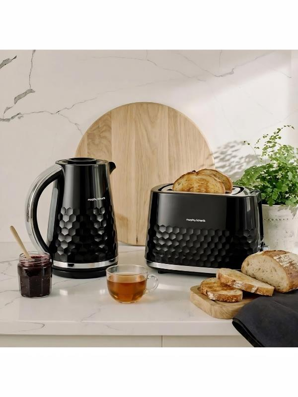Tezgahta siyah su ısıtıcısı, tost makinesi, reçel, çay ve ekmek dilimleri.