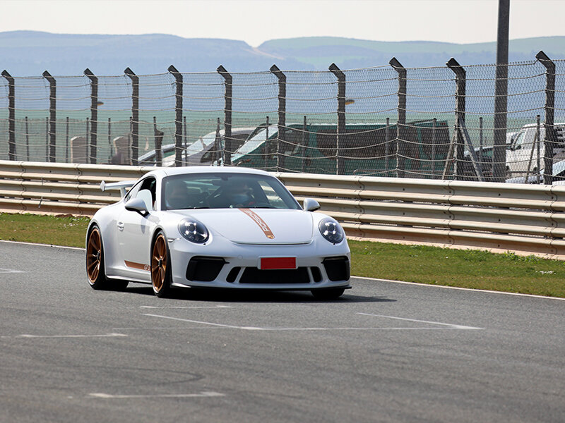 Un Porsche 911 GT3 blanco corre en una pista, con una valla y colinas al fondo.
