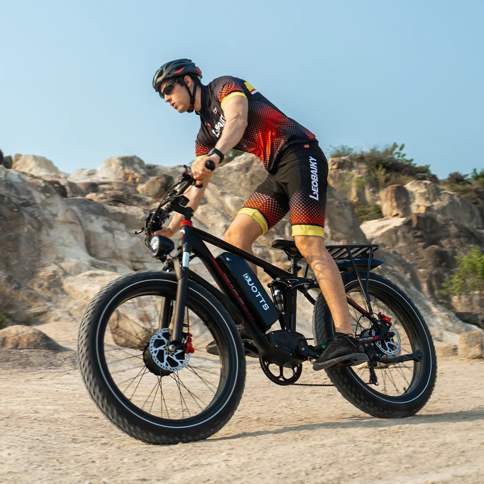 Hombre con ropa de ciclismo monta una e-bike negra de llantas anchas en sendero rocoso.