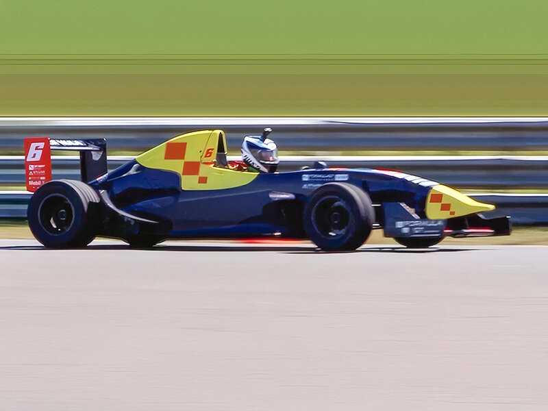 Un coche de carreras azul y amarillo, número 6, en una pista. Verde y gris al fondo.