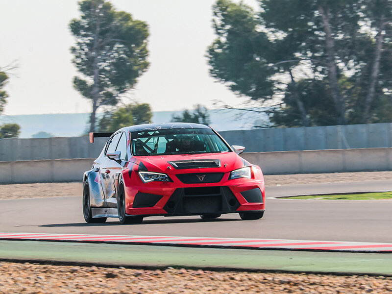 Un coche de carreras rojo en una pista con árboles y una valla al fondo.