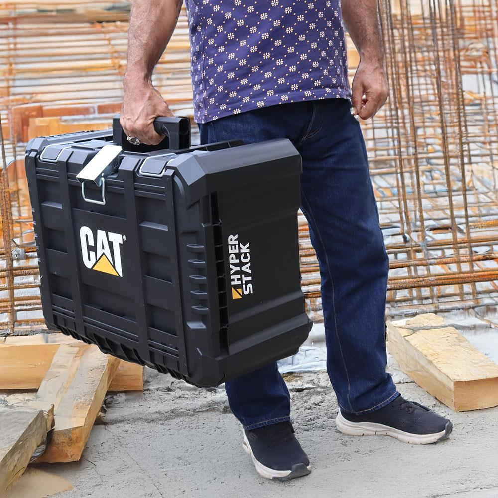 Adam inşaat alanında sarı logolu siyah CAT alet kutusu taşıyor.