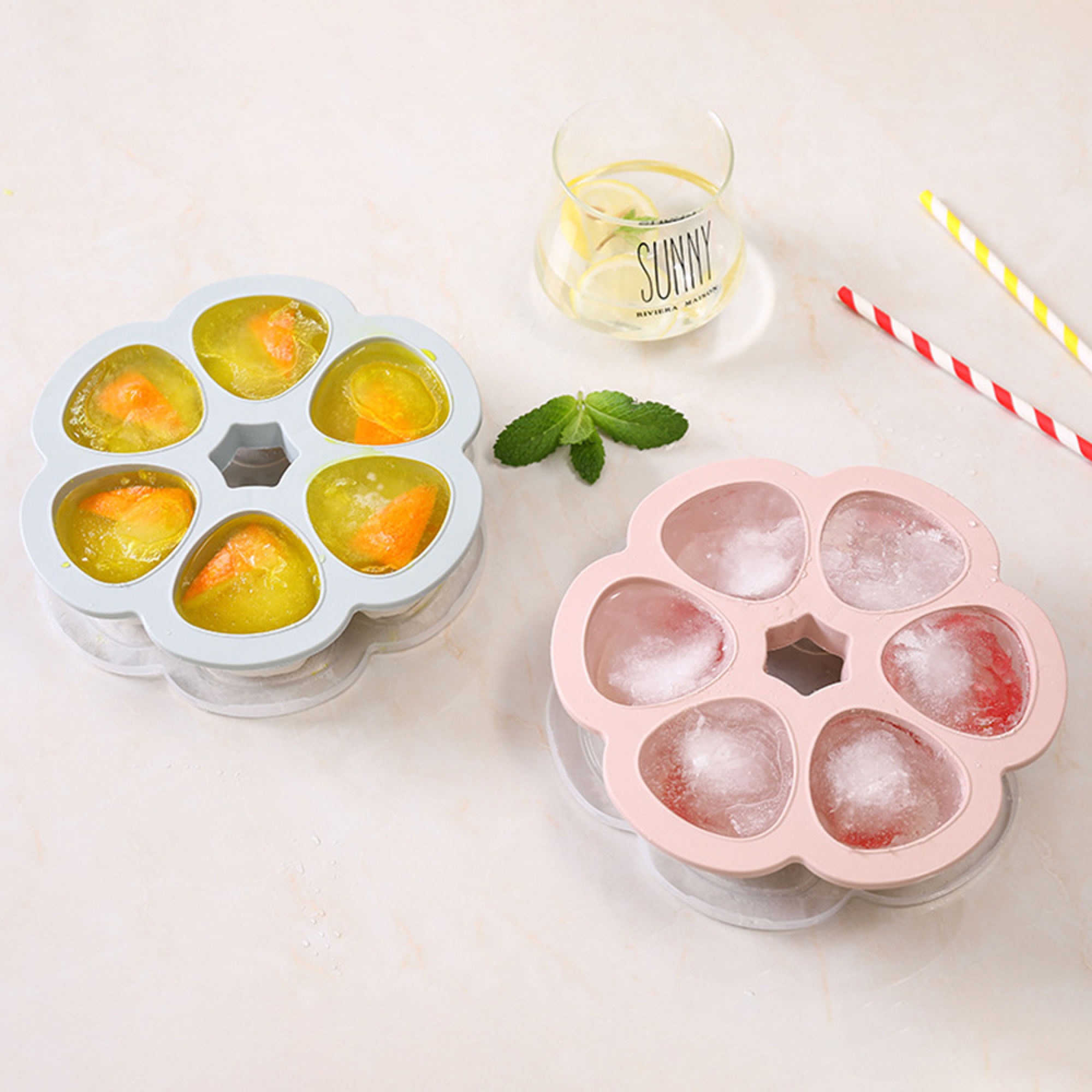 Zwei Blumenförmige Eiswürfelformen, eine hellblau mit gefrorenem Saft, eine rosa mit Eis.