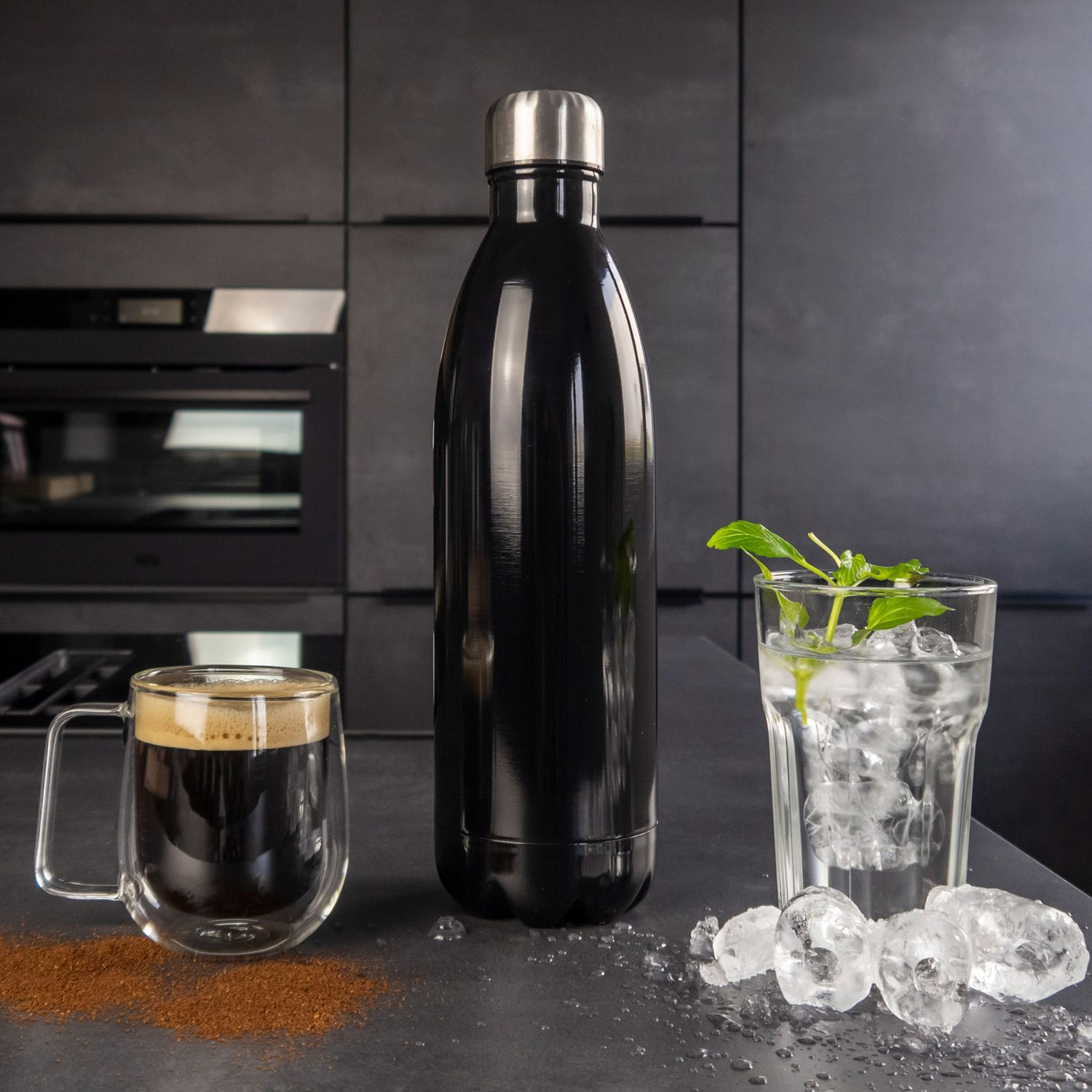 Schwarze Thermoskanne neben Kaffee und Eiswasser mit Minze auf dunkler Arbeitsplatte.
