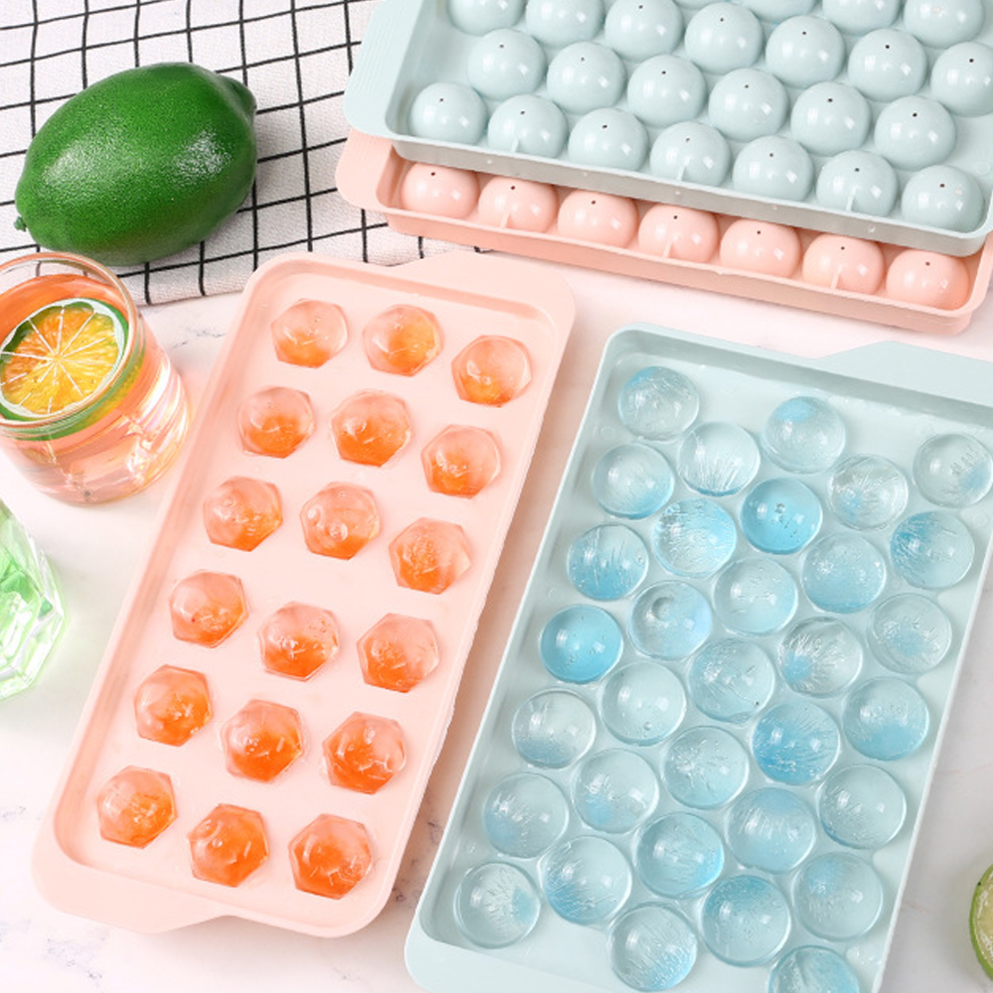 Zwei Eiswürfelformen mit runden Eiswürfeln. Eine Form hat orange, die andere blaue Eiswürfel. Eine Limette und ein Getränk sind in der Nähe.