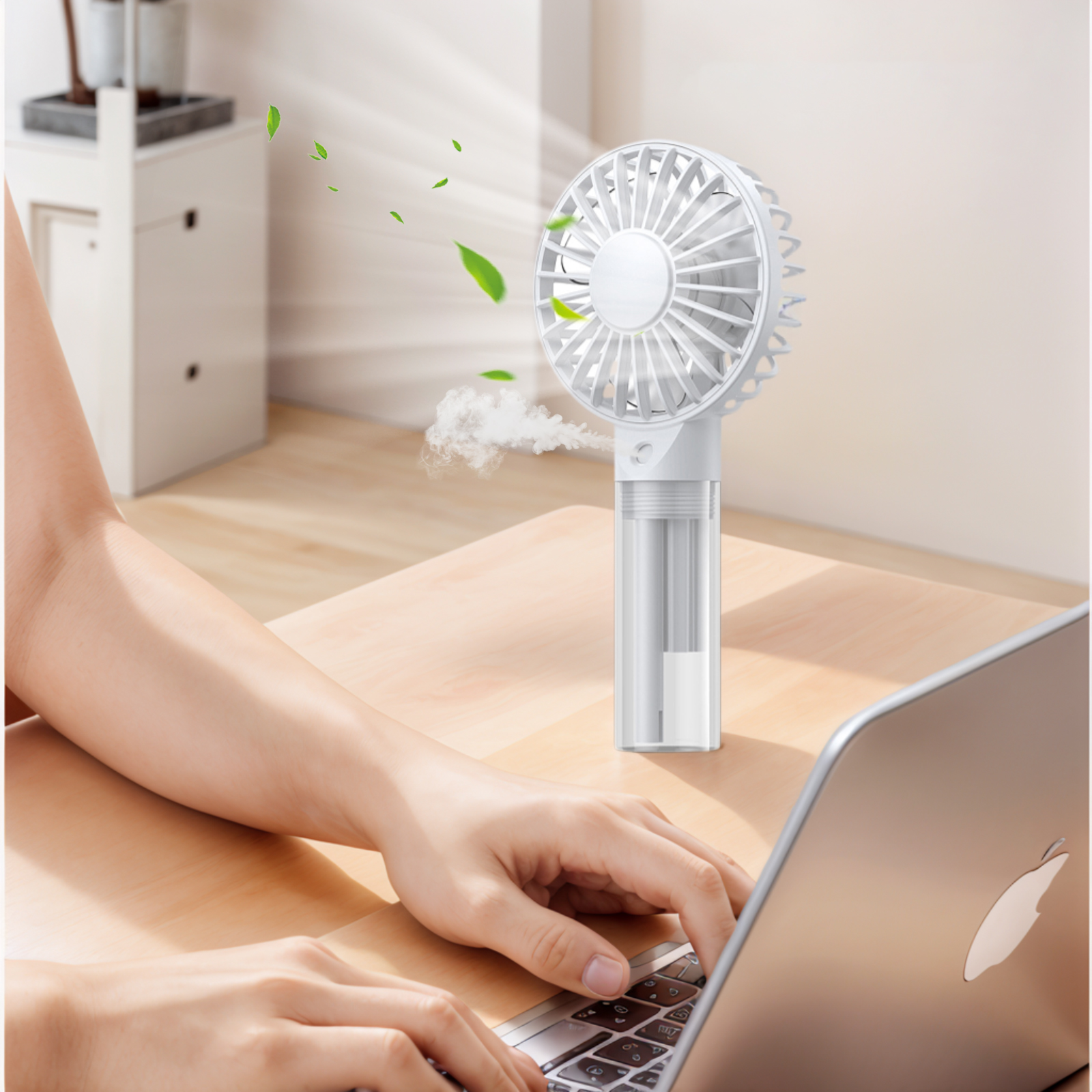 Ein weißer Handventilator sprüht Nebel, Blätter fliegen. Eine Person tippt auf einem Laptop.