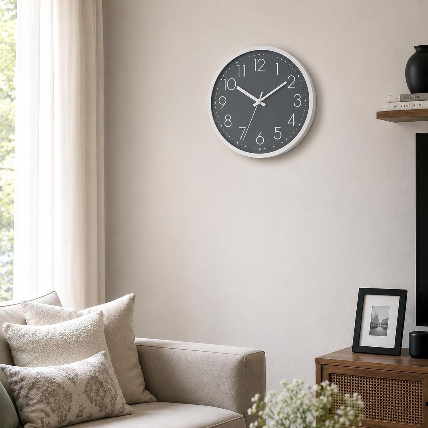 Eine graue Wanduhr mit weißen Ziffern und Zeigern hängt an einer Wand neben einem Fenster.