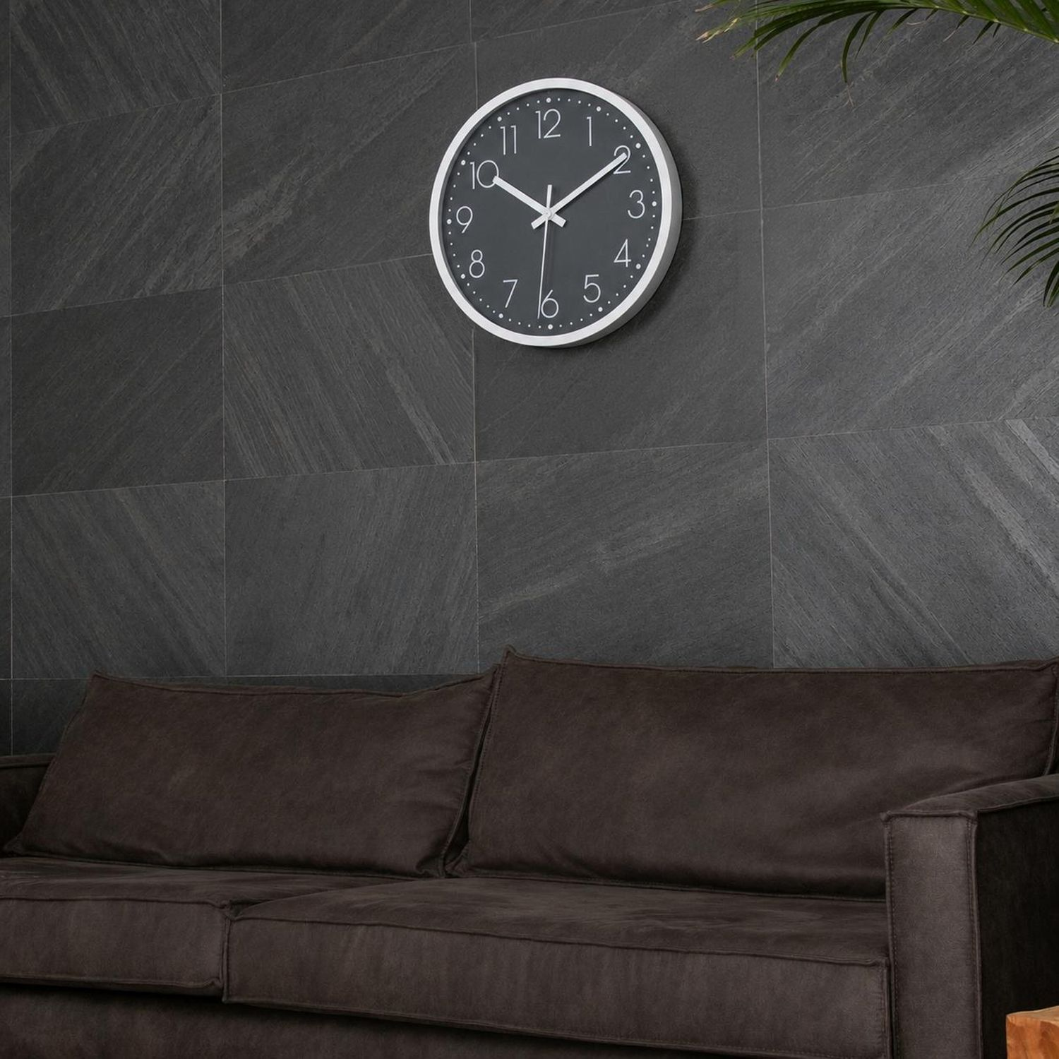 Eine dunkelgraue Wanduhr mit weißen Zeigern hängt über einem braunen Sofa.