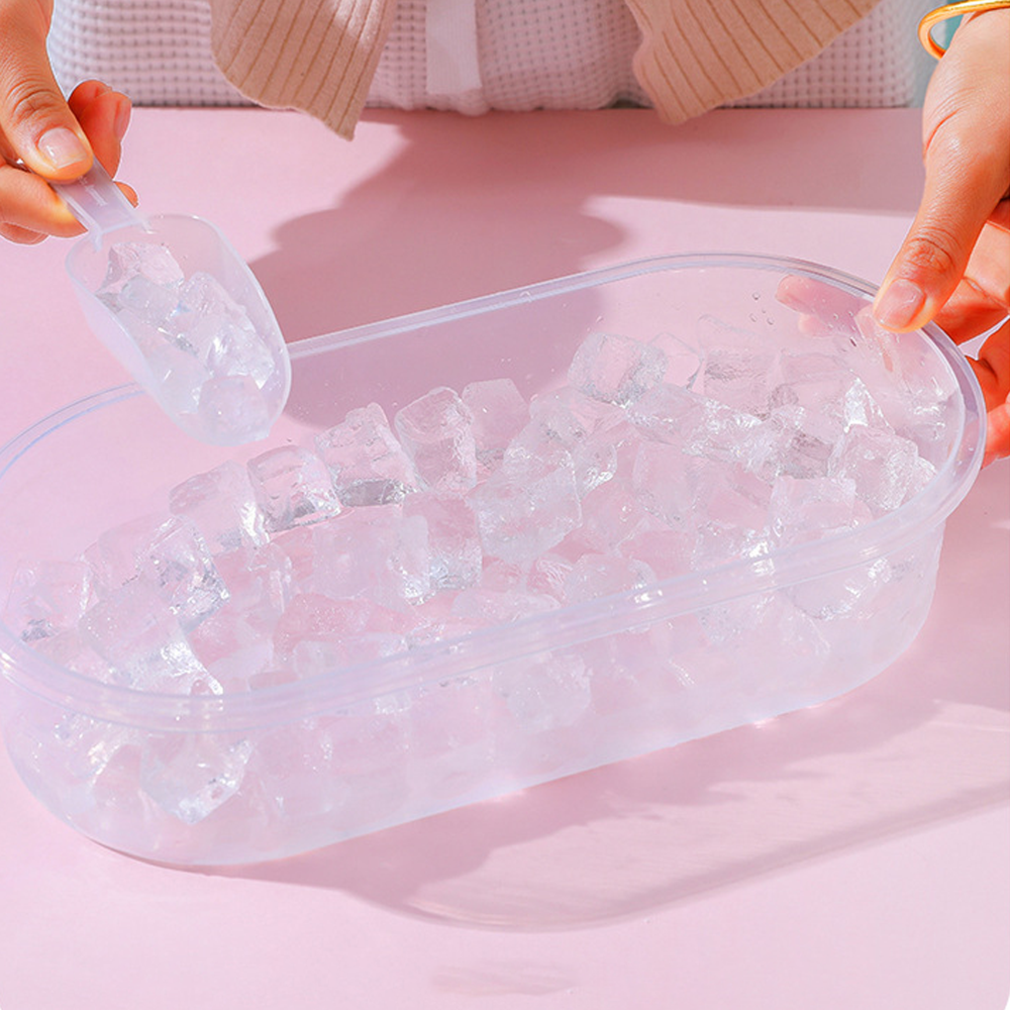 Hände schaufeln Eiswürfel in einen durchsichtigen Plastikbehälter auf rosa Untergrund.