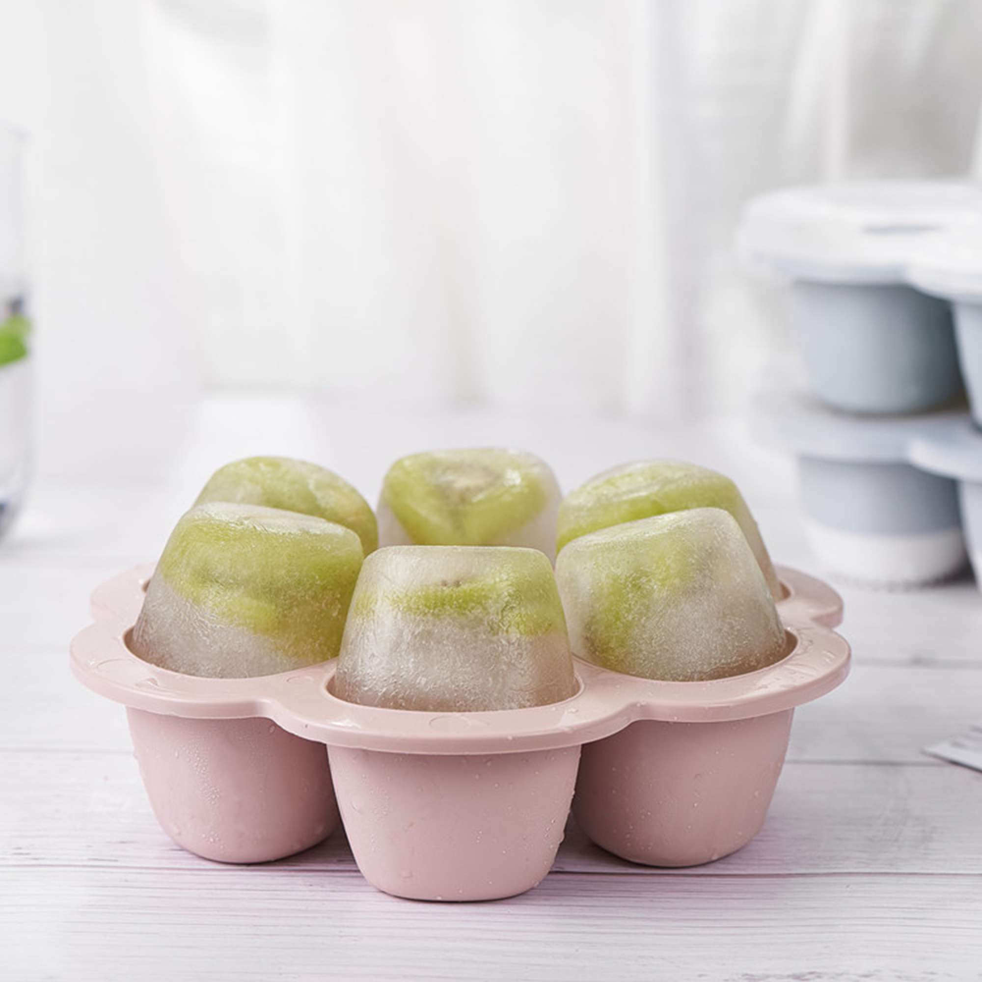 Sechs Eiswürfel mit grünen Fruchtstücken in einer rosafarbenen Form auf einem Holztisch.