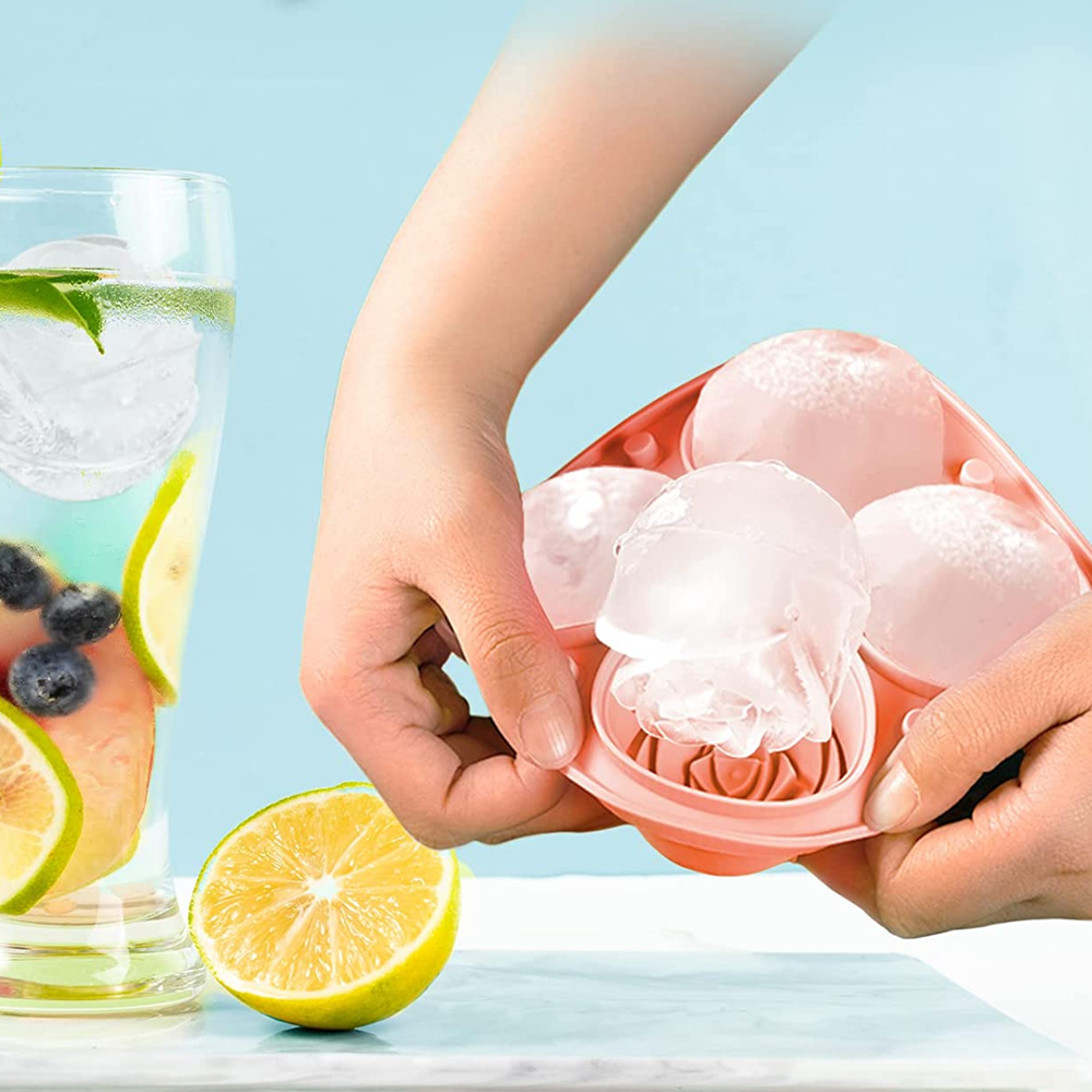 Hände halten eine rosafarbene Eiswürfelform mit runden Eiswürfeln. Daneben ein Glas mit Getränk.