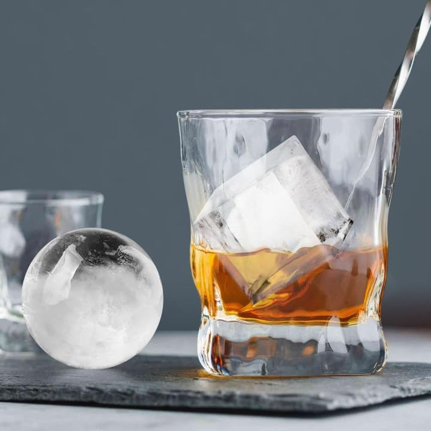 Ein Glas Whiskey mit Eiswürfeln und ein runder Eiswürfel daneben.