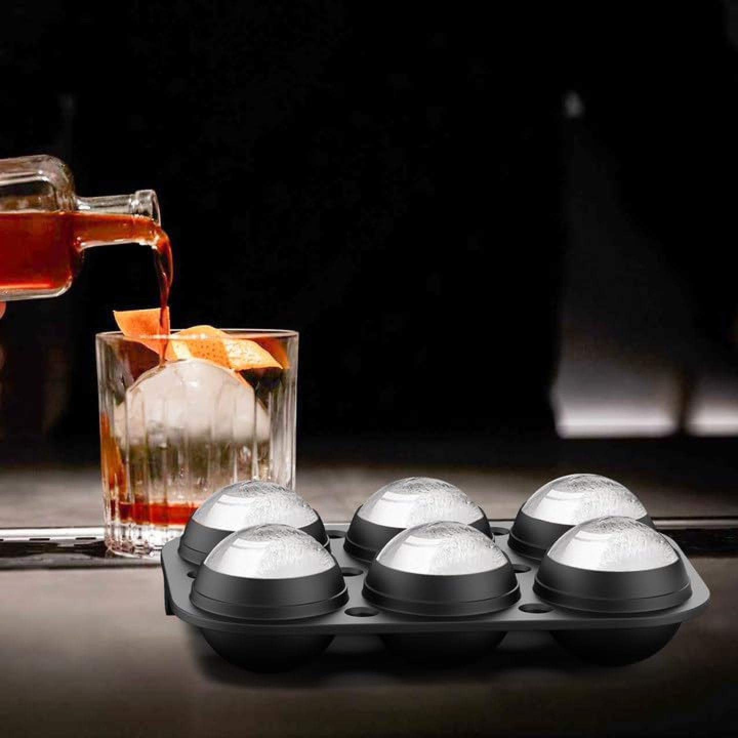 Ein Cocktail wird in ein Glas mit einem großen Eisball gegossen. Eine Eisform ist im Vordergrund.