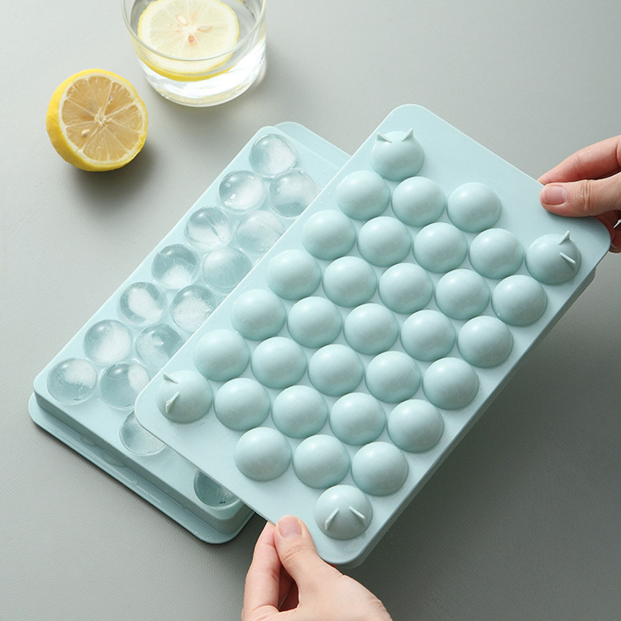 Zwei hellblaue Eiswürfelformen, eine mit Eiswürfeln, daneben eine halbe Zitrone und ein Glas Wasser.