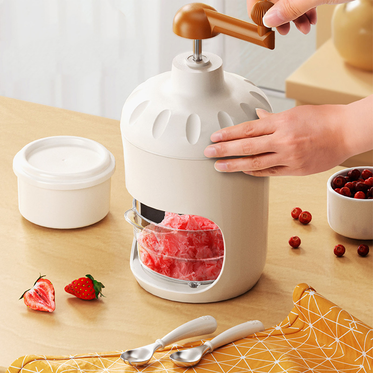 Eine Hand dreht einen Griff an einer Eismaschine mit zerstoßenem Eis. Erdbeeren und Becher sind daneben.