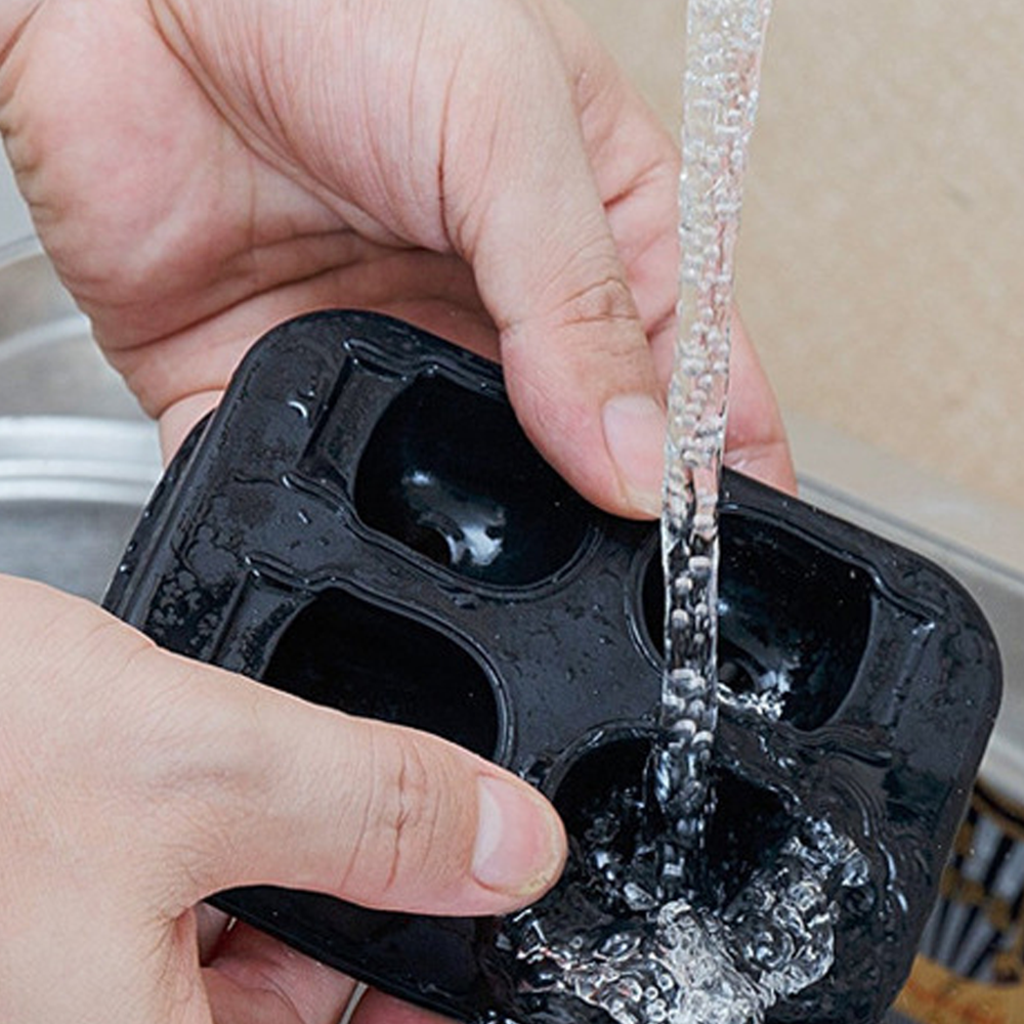 Hände halten eine schwarze Eiswürfelform unter fließendes Wasser.