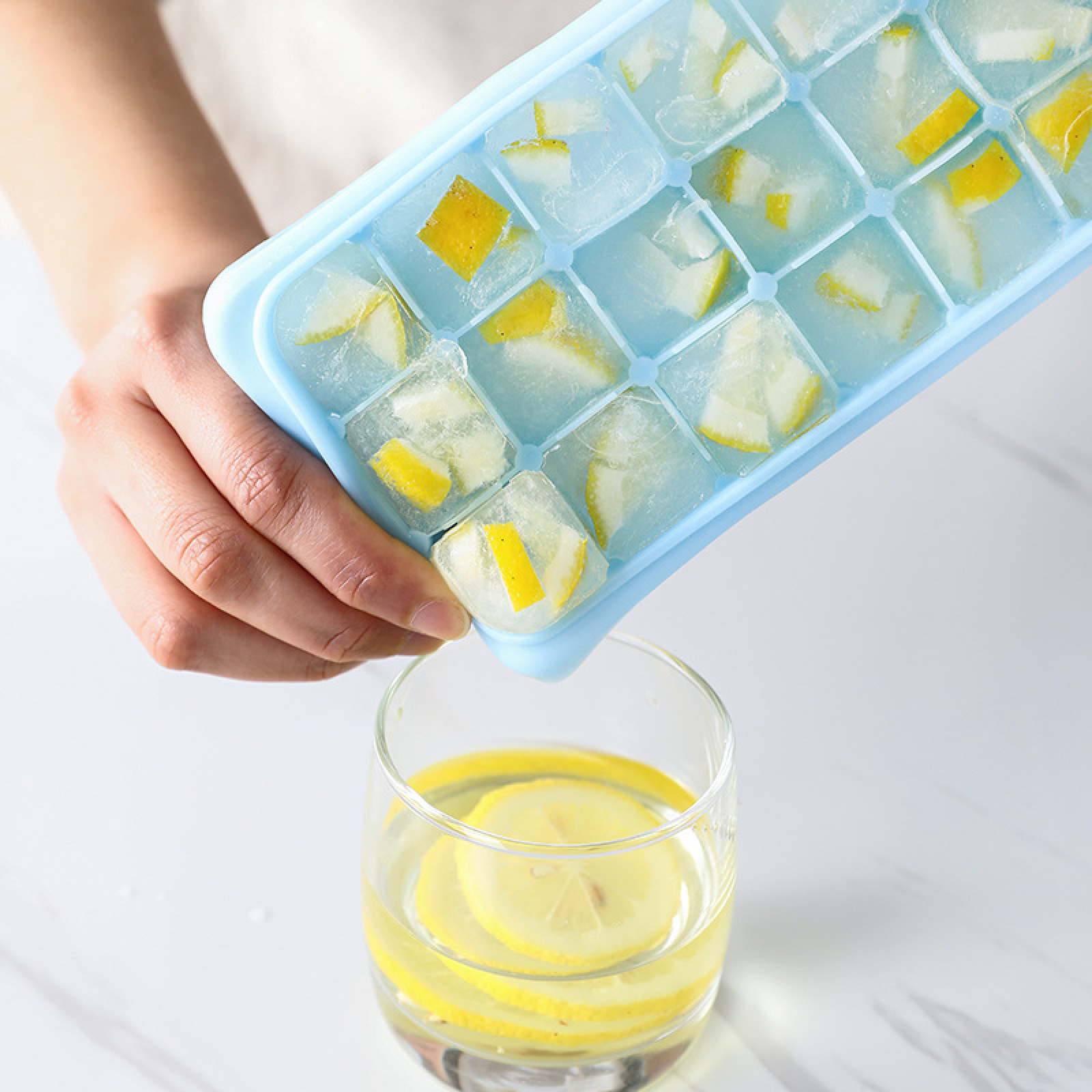 Eine Hand hält eine blaue Eiswürfelform mit Zitronen-Eiswürfeln über einem Glas Wasser mit Zitronenscheiben.