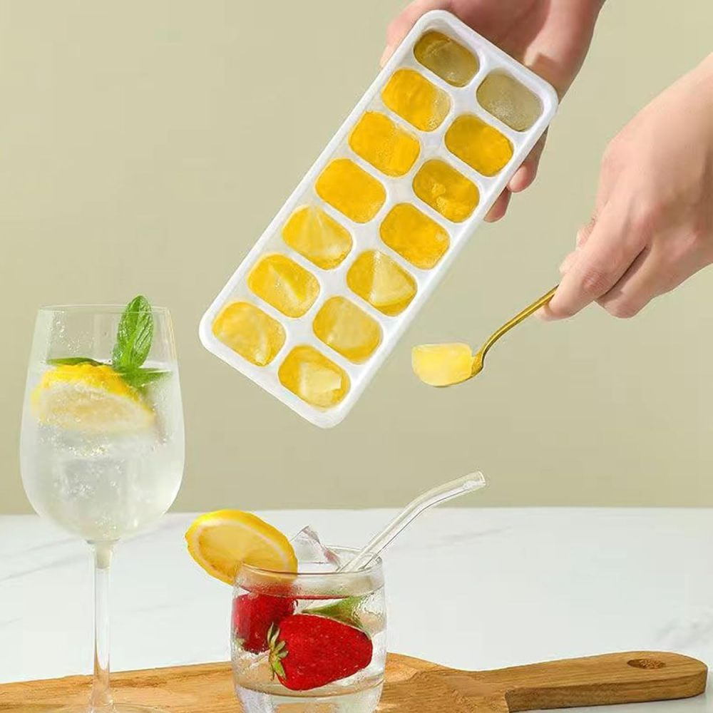 Eine Hand hält einen Löffel mit einem gelben Eiswürfel über einer weißen Eiswürfelschale mit Eiswürfeln.